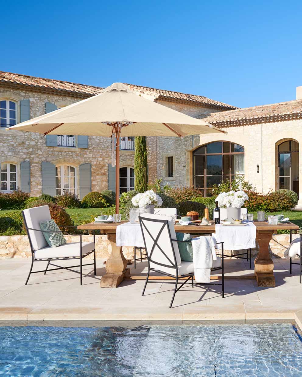 Gartenmöbel-Set mit Holztisch und beigem Sonnenschirm am Pool vor steinernem Haus