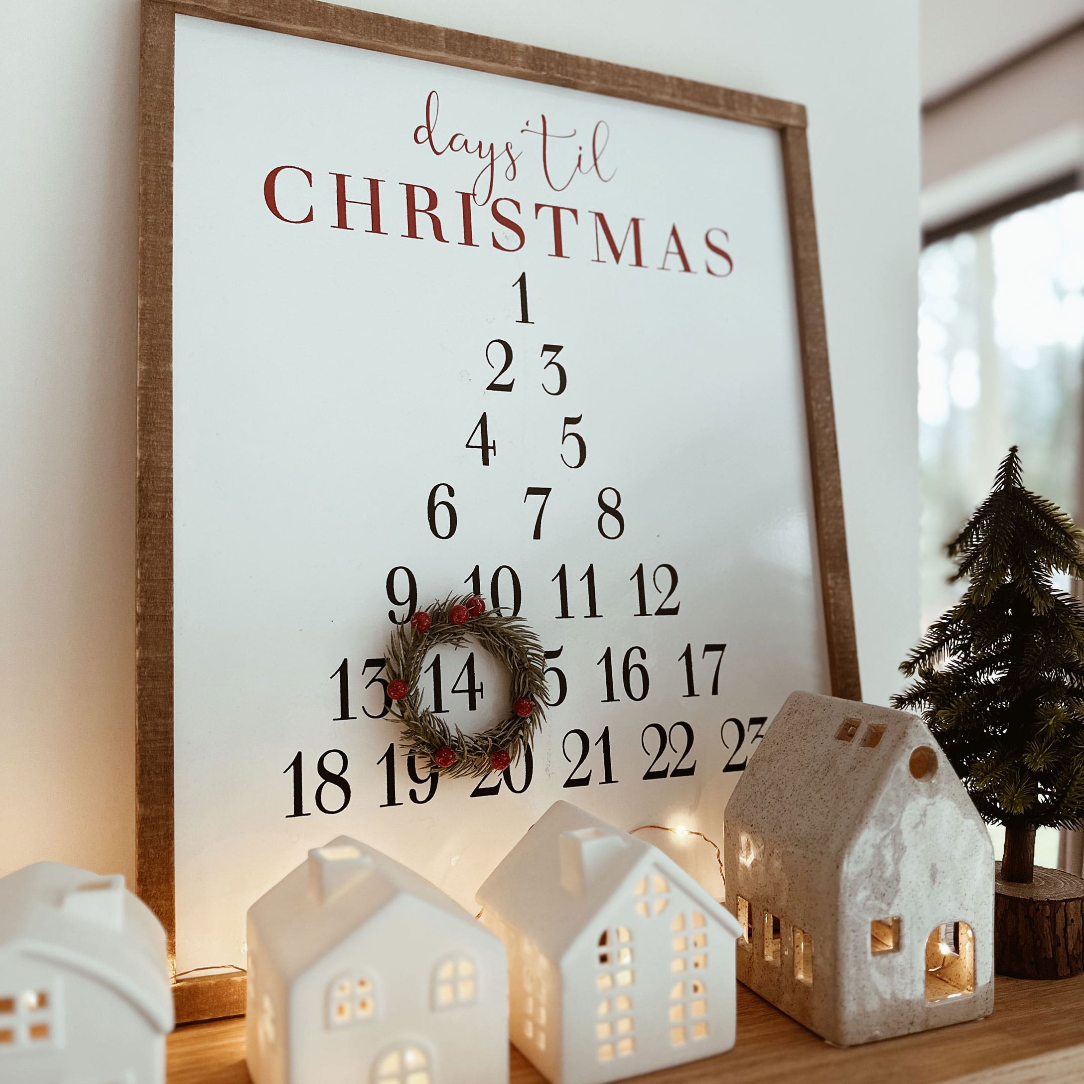 Holzrahmen mit Adventskalender und dekorativem Kranz, Keramikhäusern und Tannenbaum auf Tisch