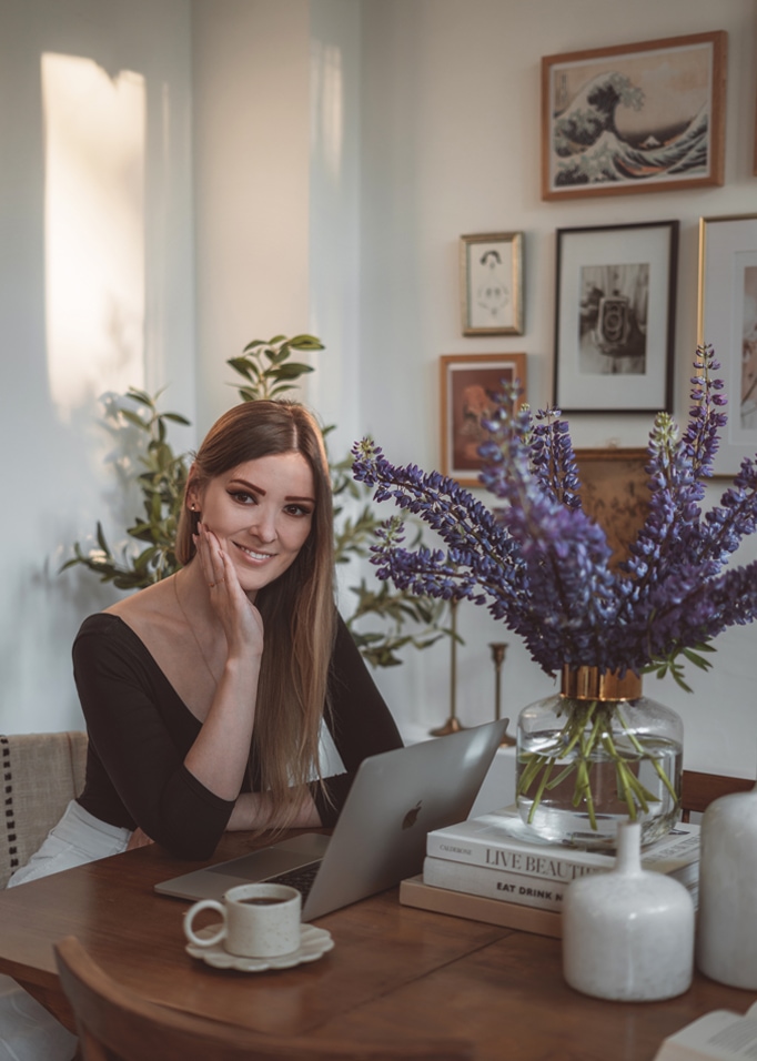 Frau am Holztisch mit Laptop und Glasvase mit violetten Blumen in hellem Raum.
