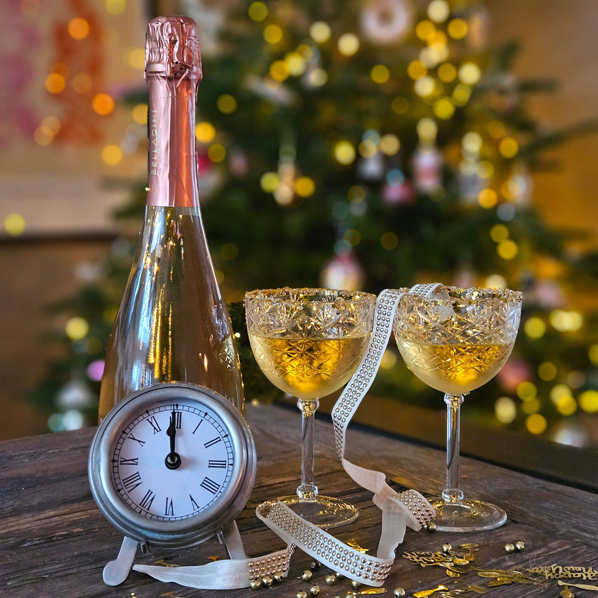 Flasche Champagner mit rosa Folie, zwei Kristallgläser mit goldenem Getränk und Uhr auf Holztisch