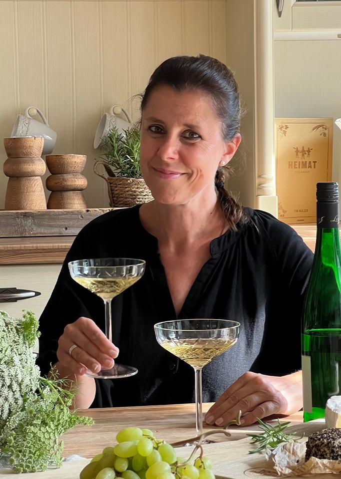 Frau in schwarzer Bluse hält ein Weissweinglas am Tisch mit Trauben und Käse.