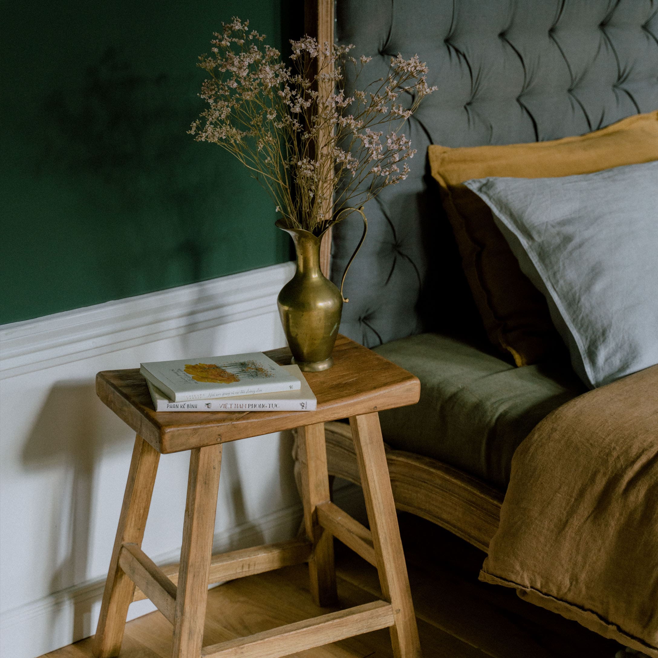 Holzhocker mit Messingkrug und Trockenblumen neben einem Bett mit grauem Polster und brauner Bettwäsche