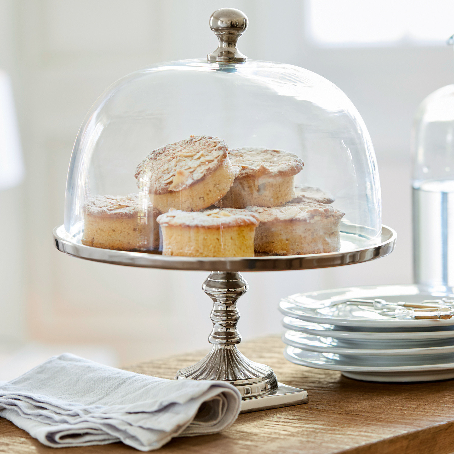 Glasglocke mit Platte Mine Metall-Tortenplatte mit transparenter Glasglocke und Kuchen auf Holztisch neben weissen Tellern
