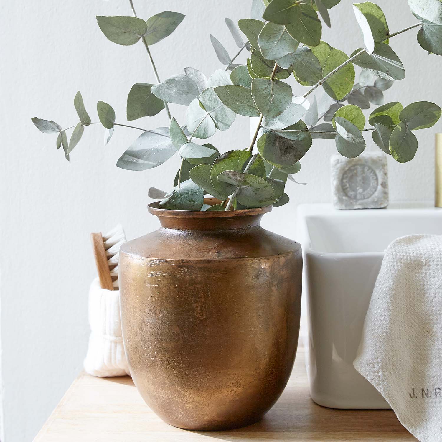 Kupferfarbener Vase mit grünen Eukalyptusblättern auf Holzplatte in hellem Raum