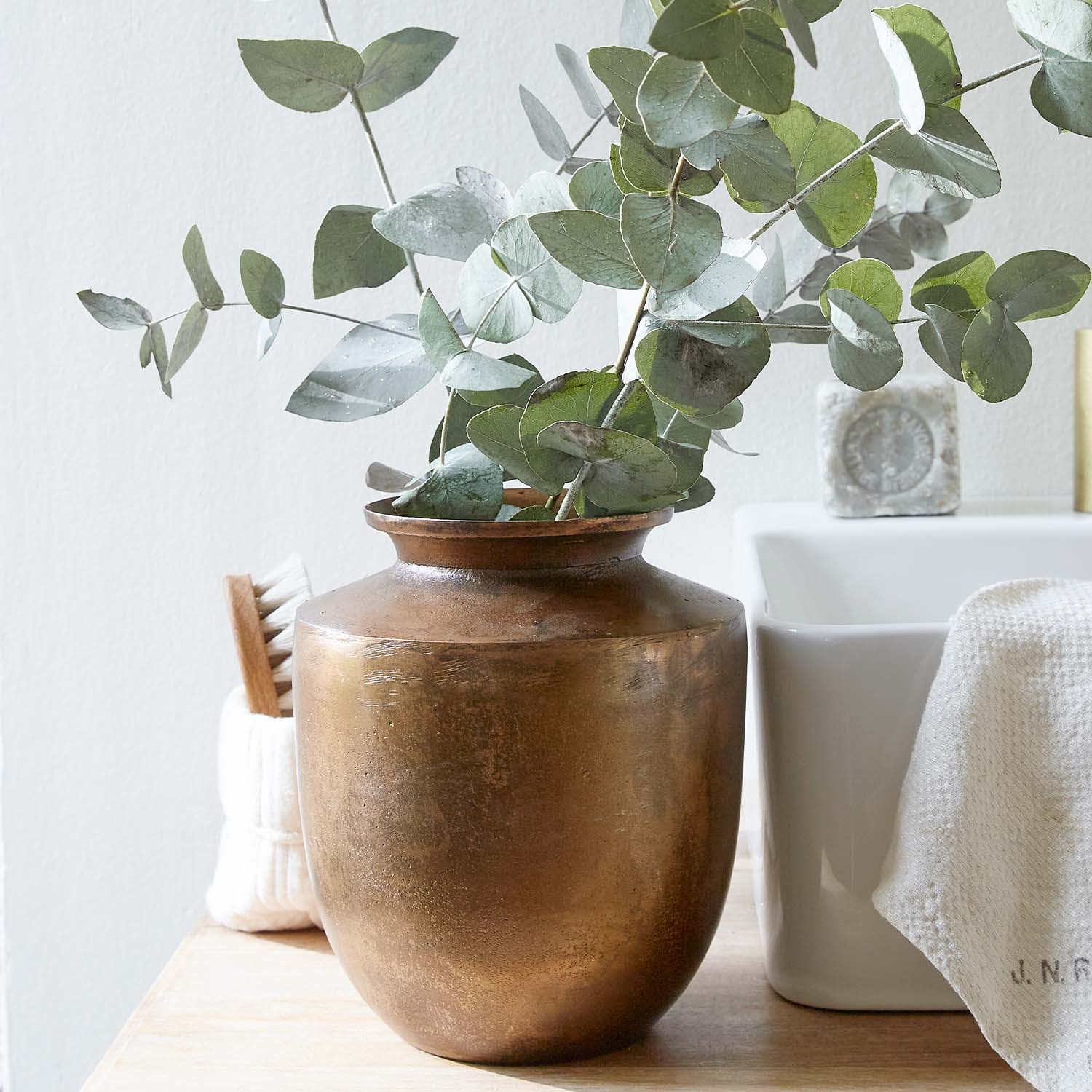 Kupferfarbener Vase mit grünen Eukalyptusblättern auf Holzplatte in hellem Raum