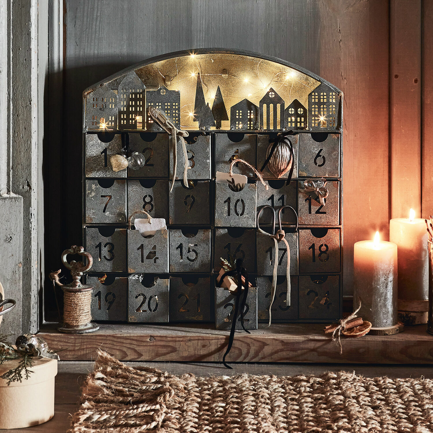 Adventskalender Léran Metall-Adventskalender mit 24 nummerierten Schubladen und beleuchteter Häusersilhouette, dekoriert mit Kerzen.