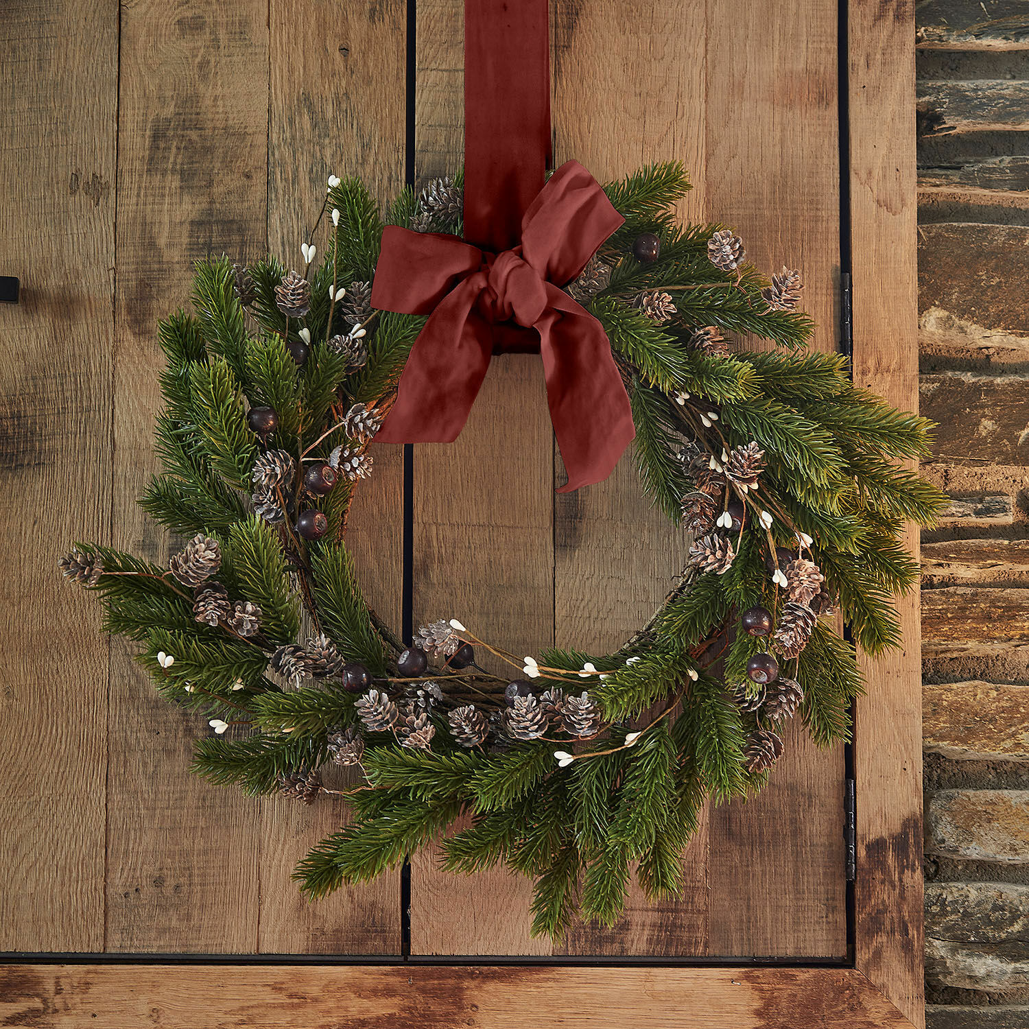 Kranz Lyandra Weihnachtskranz aus grünen Zweigen und Zapfen mit roter Schleife an Holztür