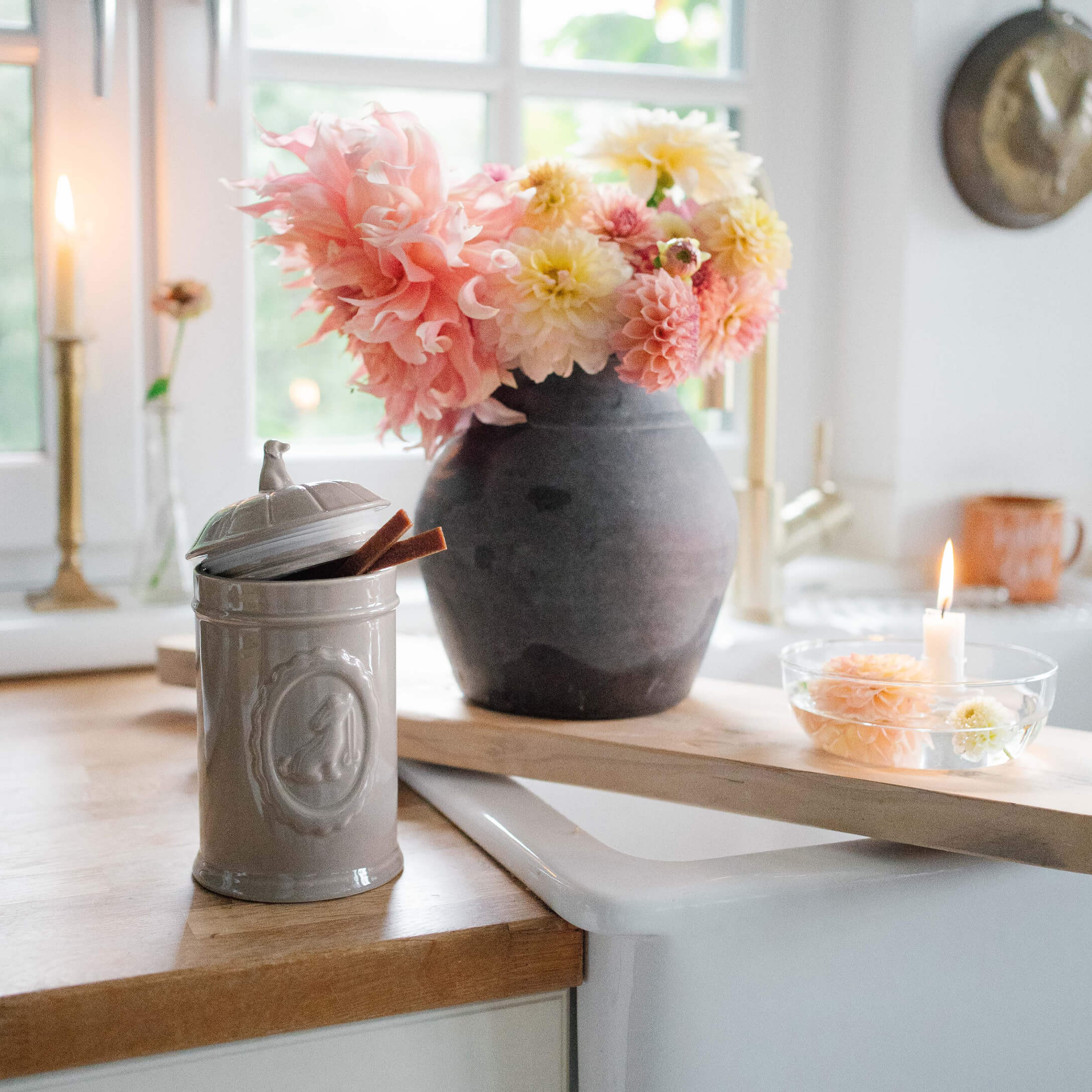 Grauer Keramikbehälter mit Deckel auf Holzarbeitsfläche neben schwarzer Vase mit rosa-gelben Blumen