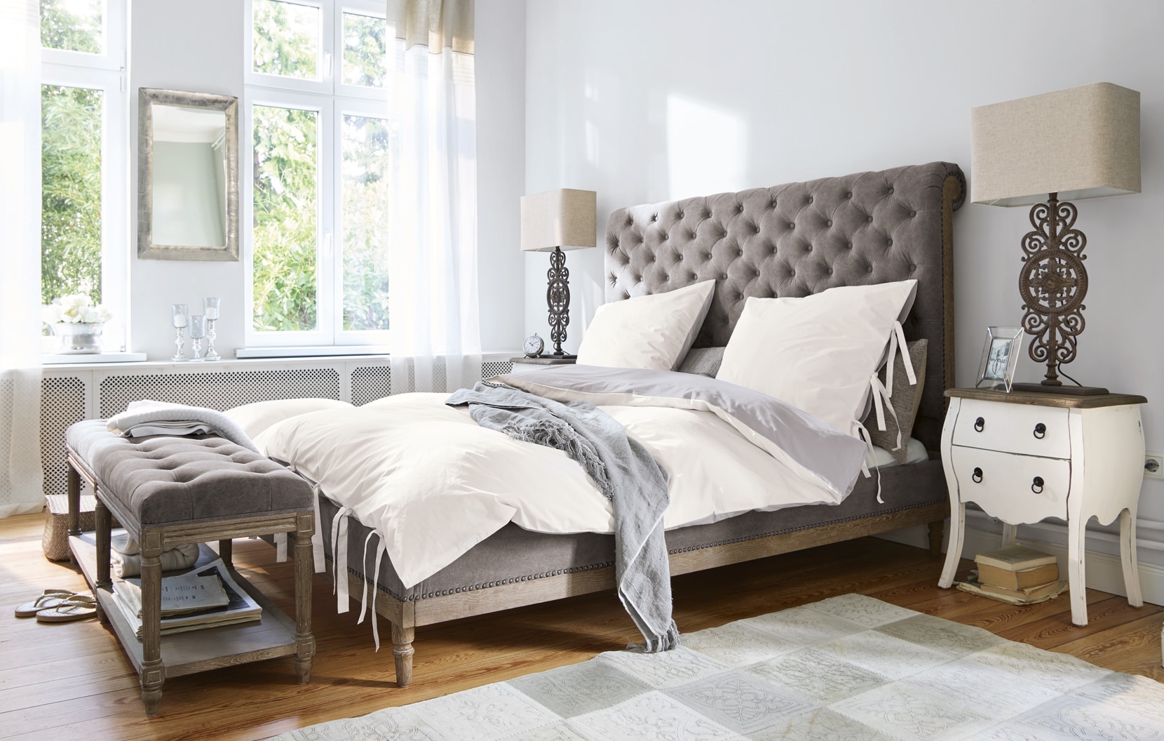 Klassisches Schlafzimmer mit grauem Polsterbett und weisser Bettwäsche in hellem Raum