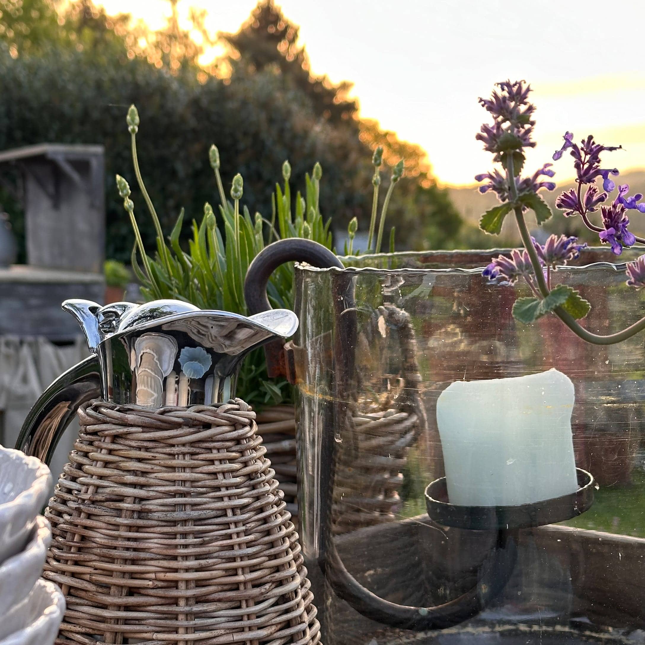 Metallkrug mit Korbgeflecht und Glas-Kerzenhalter im sommerlichen Garten