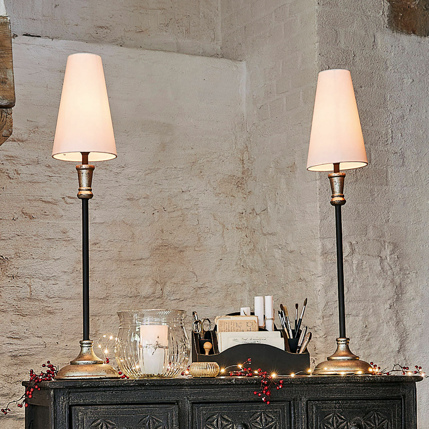 Tischlampe 2er Set Obsonville Ein Paar Tischlampen mit Metallfuss und weissen Lampenschirmen auf einem dunklen, verzierten Sideboard.