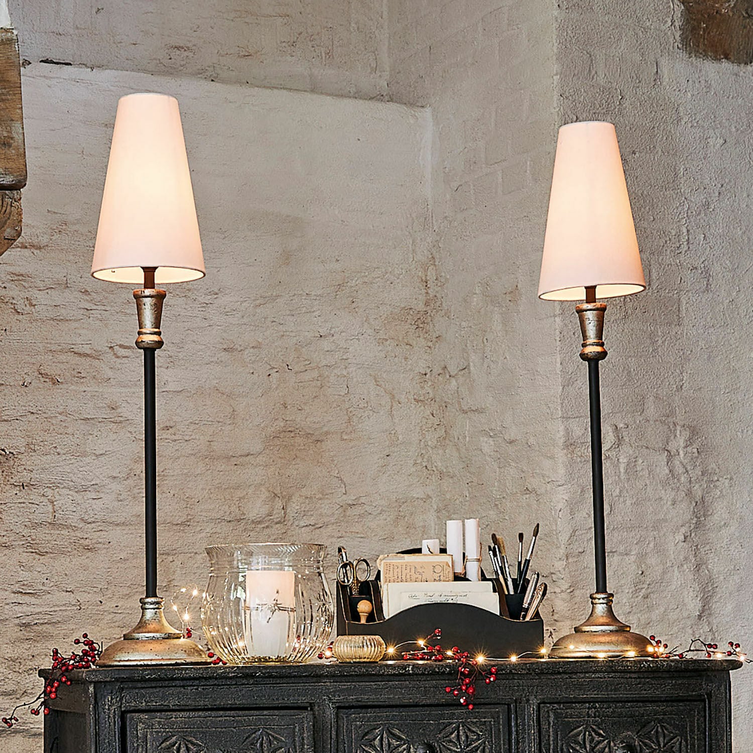 Tischlampe 2er Set Obsonville Ein Paar Tischlampen mit Metallfuss und weissen Lampenschirmen auf einem dunklen, verzierten Sideboard.