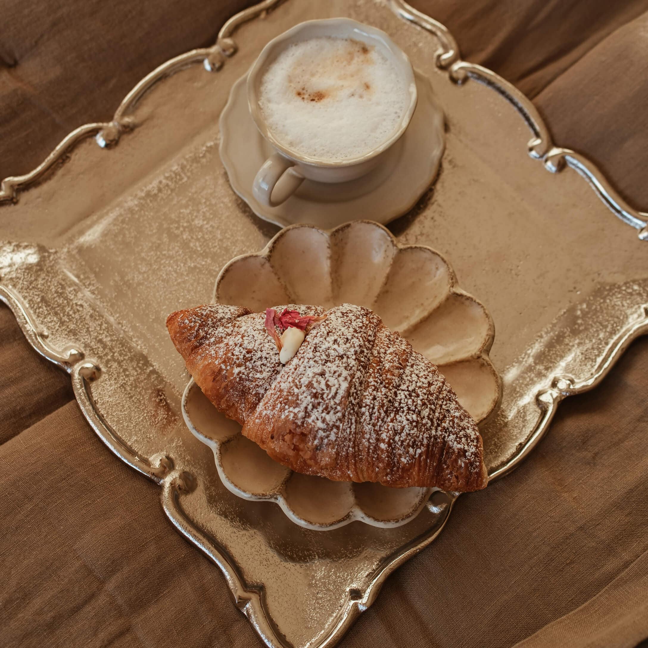 Becher cremiger Cappuccino und Croissant mit Puderzucker auf beigem Teller und goldfarbenem Tablett