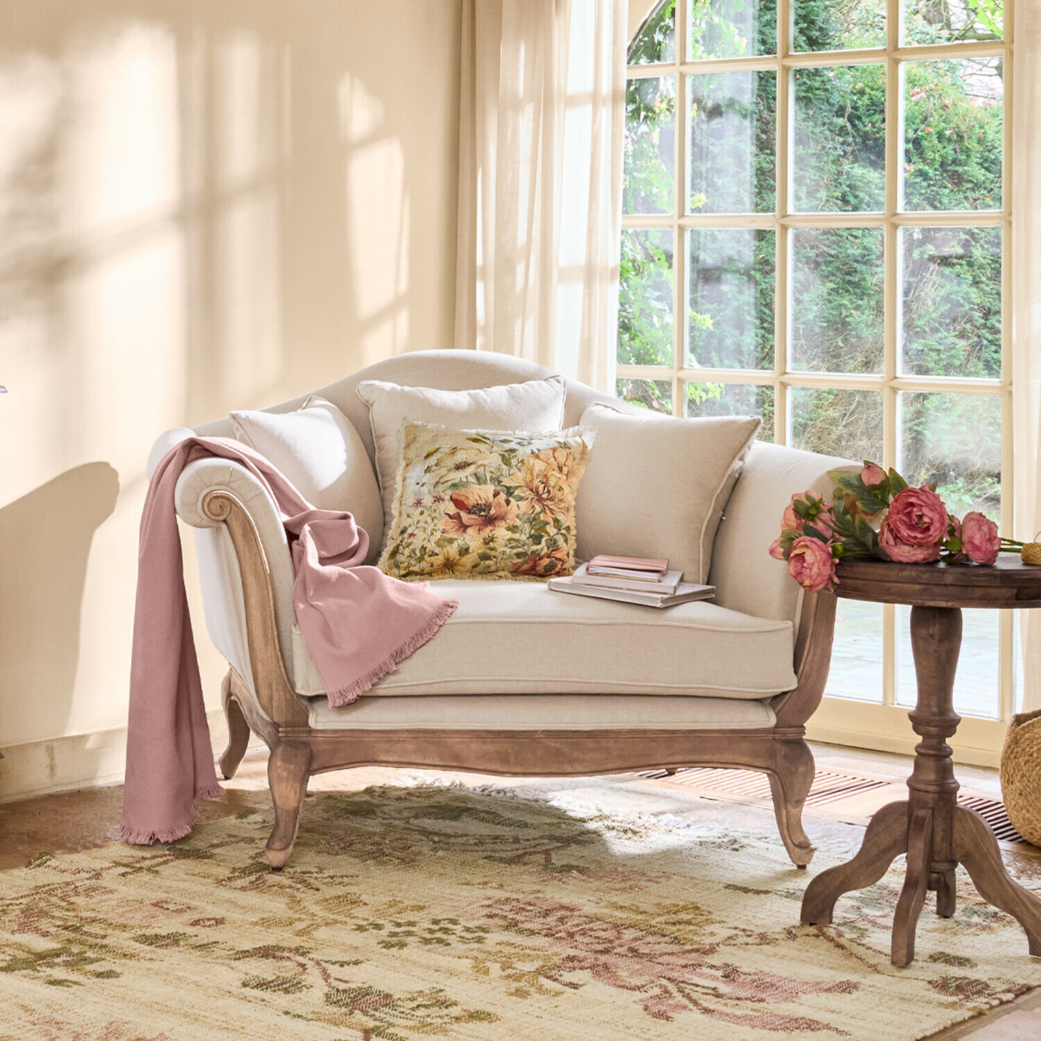 Beiges Sofa mit Holzrahmen, floralem Kissen und rosa Decke in hellem Wohnzimmer mit Fenster