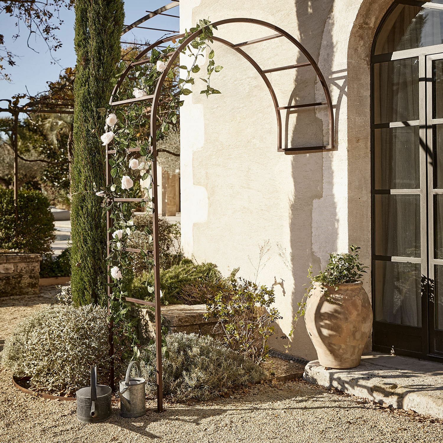 Rosenbogen Auburn Braune Metall-Gartenpergola mit Bogen, weissen Rosen und Gieskannen auf Kiesboden