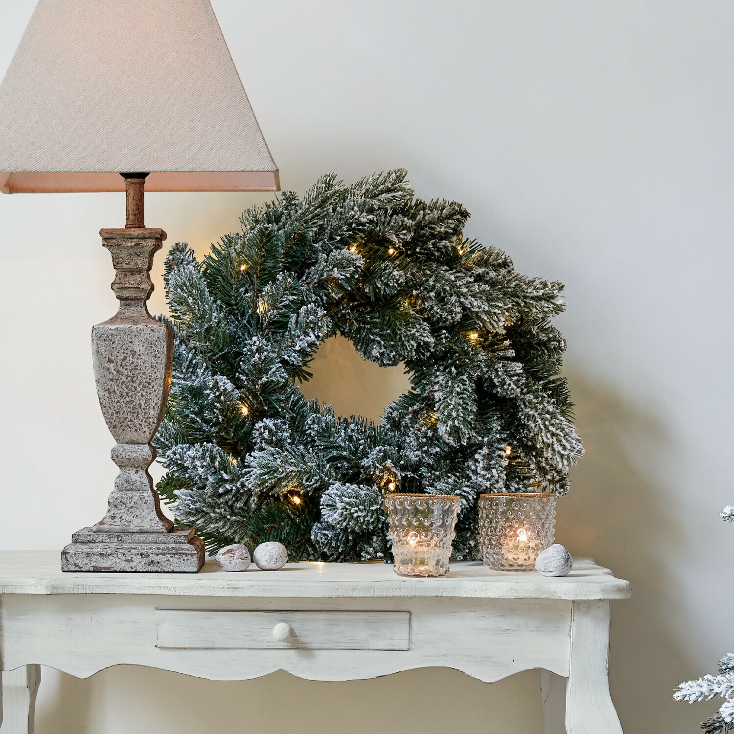 LED-Kranz Regin Schneebedeckter Weihnachtskranz mit grünen Zweigen und Lichterkette auf weissem Tisch mit Lampe