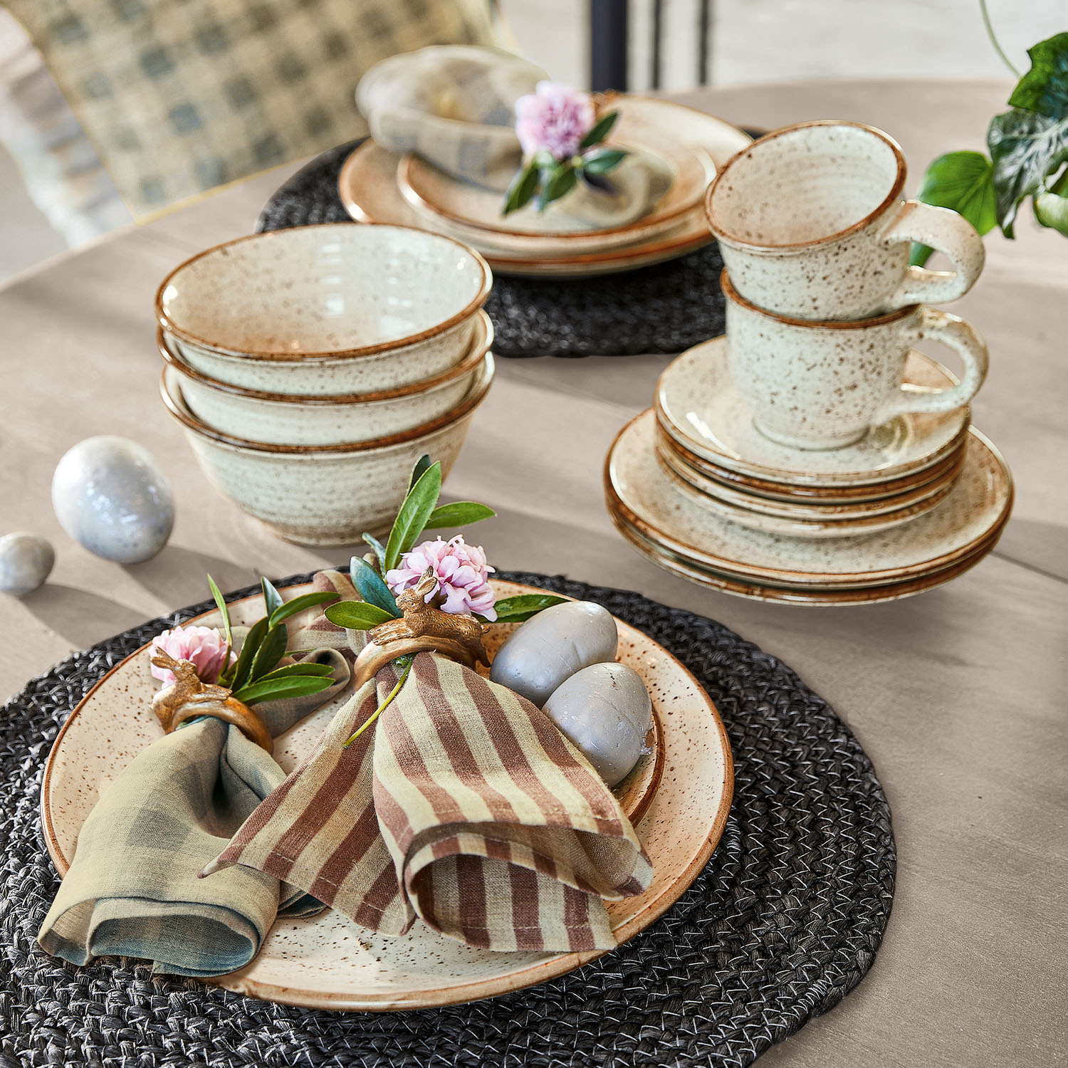 Keramik-Geschirrset in Creme mit braunen Sprenkeln und Serviettenringen mit Blumen auf schwarzem Tischset