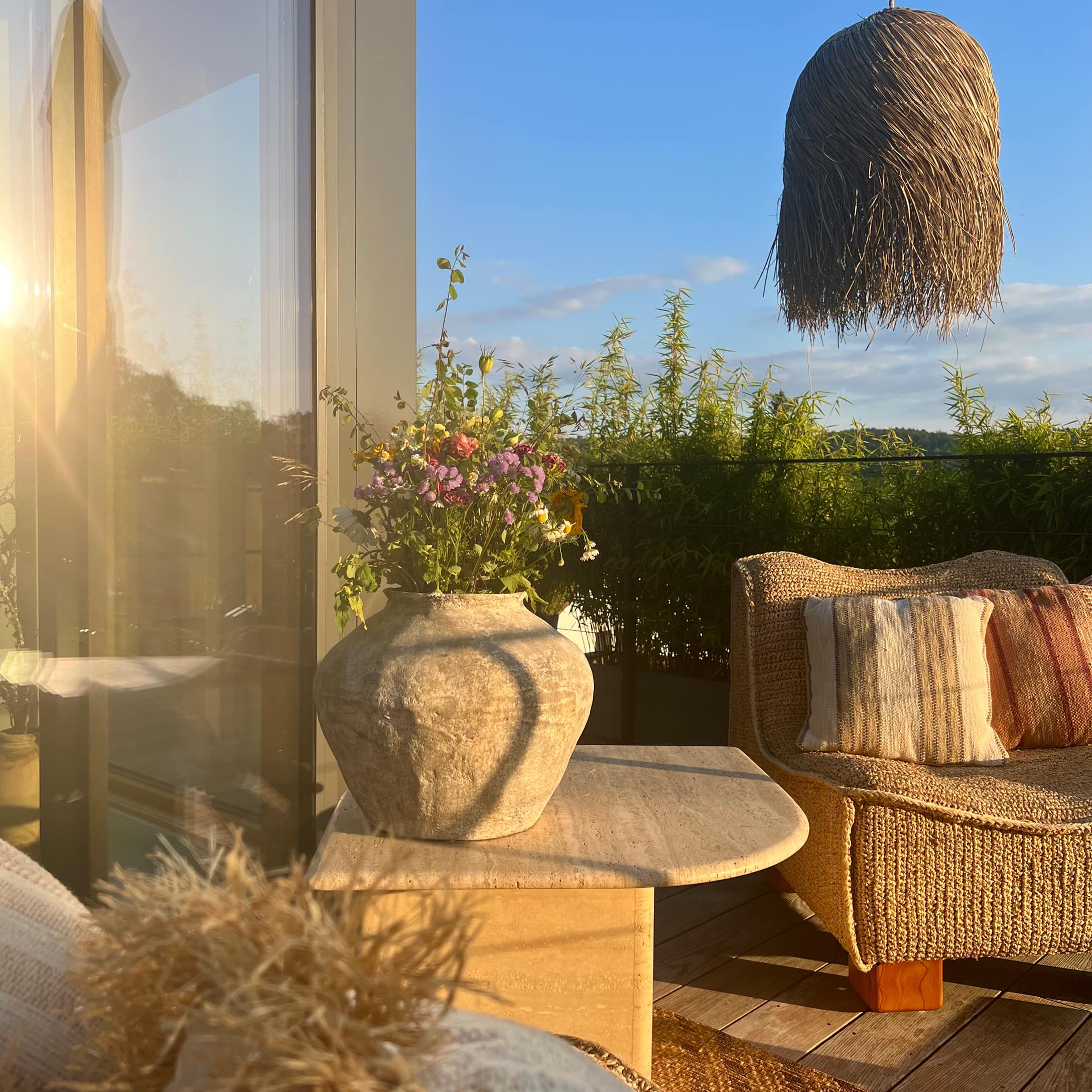 Grosse Keramikvase mit Blumen auf Steintisch auf Terrasse mit Rattansofa und Graslampe