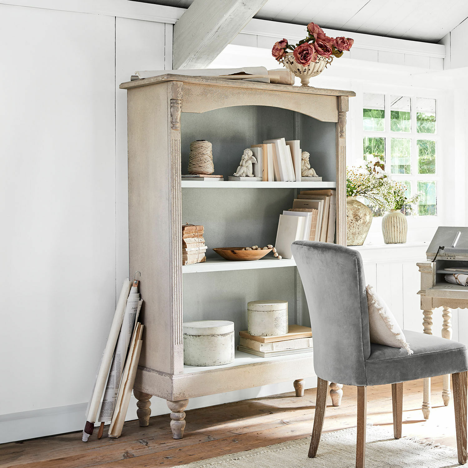 Regal Arterton Beige Holzvitrine im Vintage-Stil mit dekorativen Büchern und grauem Stuhl in hellem Raum