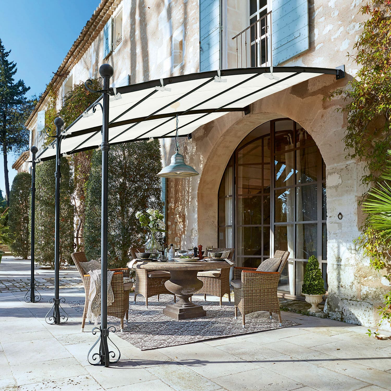 Pergola Kattles Gartenmöbel-Set aus Rattan unter weissem Vordach auf steinerner Terrasse vor einem Steinhaus