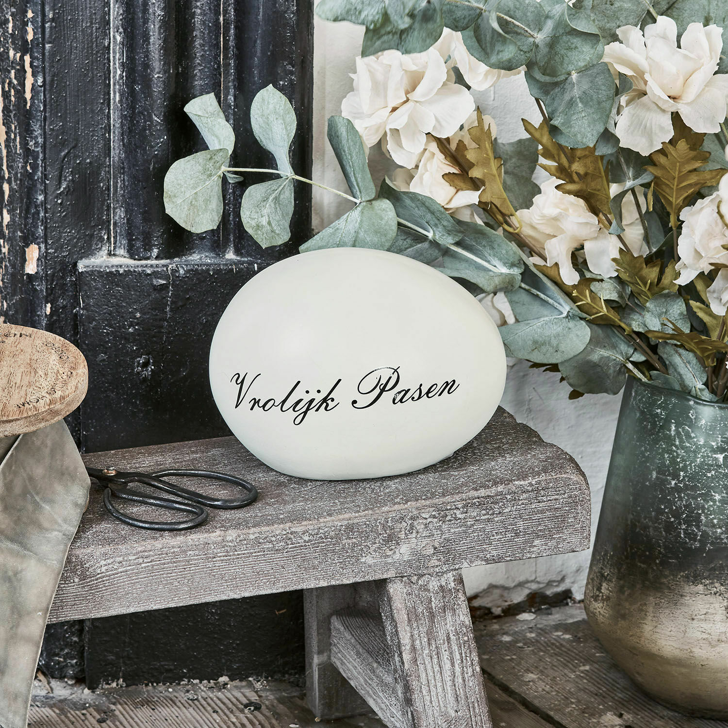 Dekoratives weisses Ei mit Schriftzug Vrolijk Pasen auf Holzbank neben Blumen und Schere