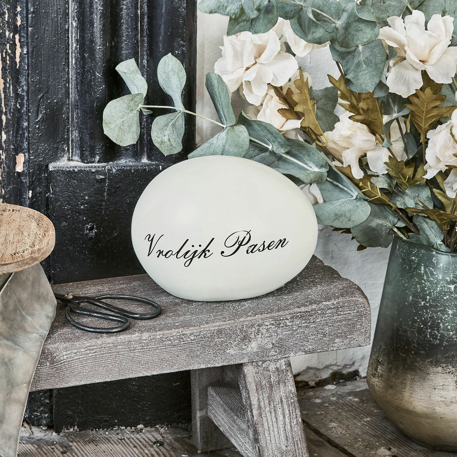 Deko-Ei VROLIJK PASEN Dekoratives weisses Ei mit Schriftzug Vrolijk Pasen auf Holzbank neben Blumen und Schere