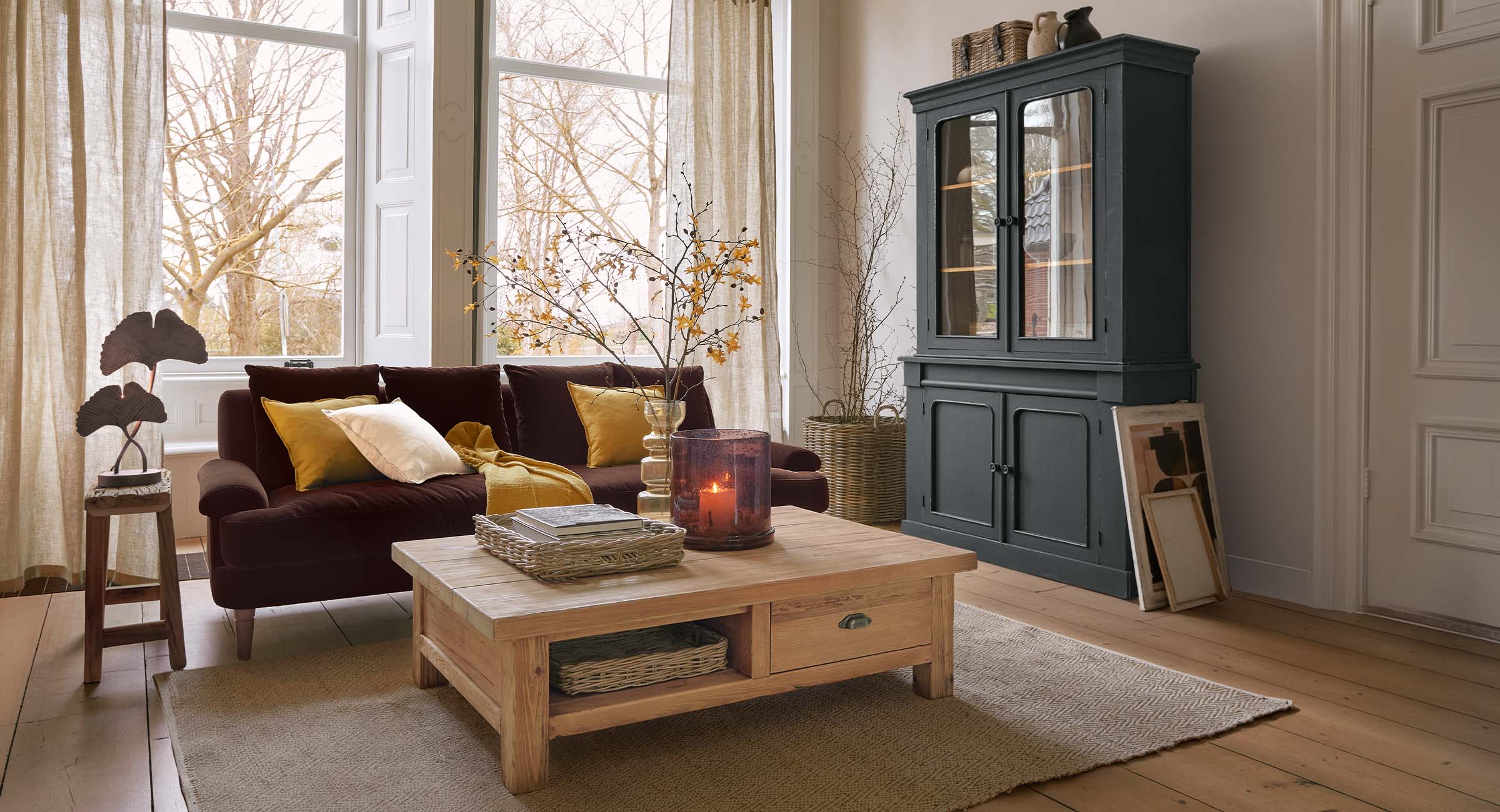 Gemütliches Wohnzimmer mit dunklem Sofa, Holztisch und schwarzem Holzvitrinenschrank vor großen Fenstern