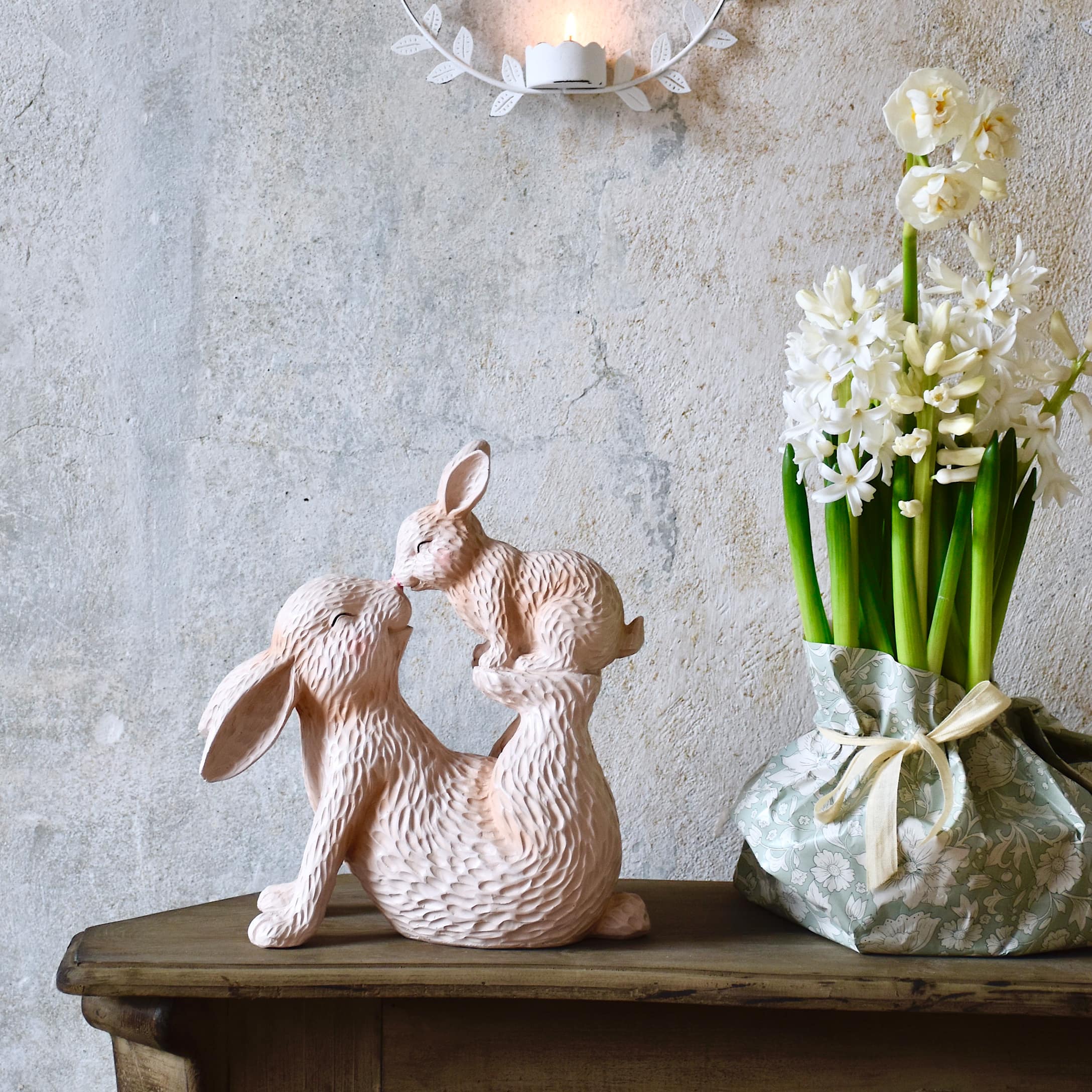 Rosa Holzskulptur mit zwei Hasen auf einem Holztisch neben Blumen in pastellfarbigem Übertopf