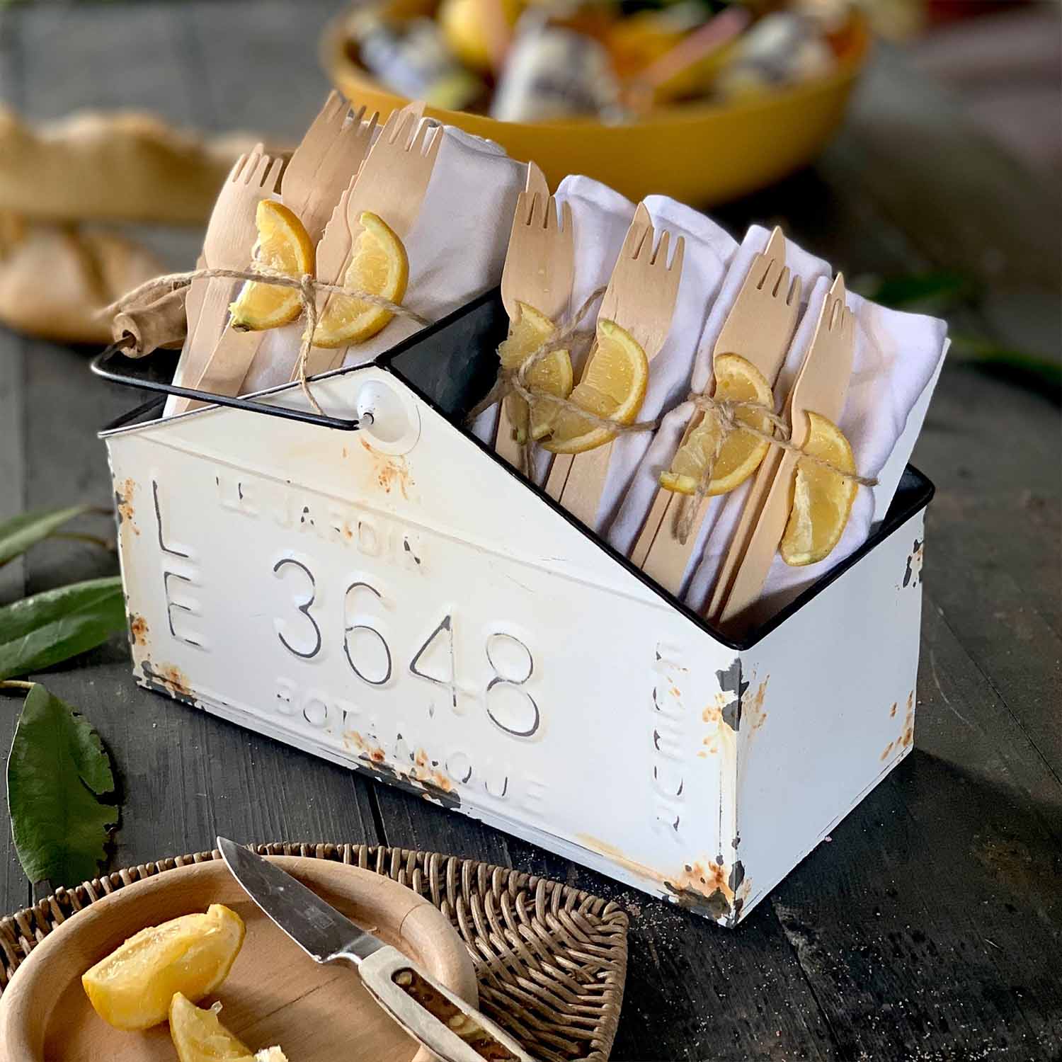Weißer Metallbehälter mit Used-Look, gefüllt mit Holzbesteck und weißen Servietten, dekoriert mit Zitronenscheiben