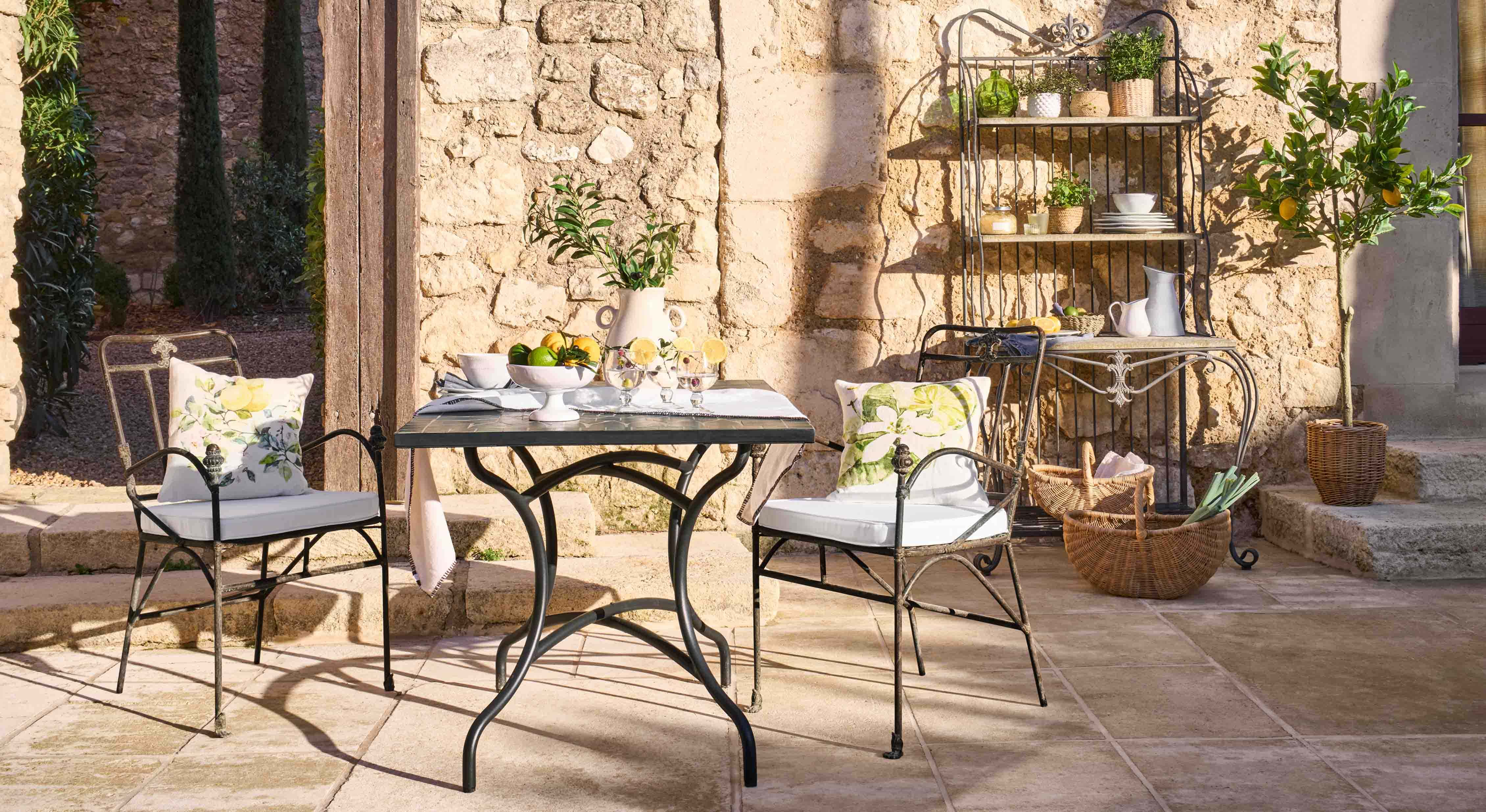 Metalltisch und Stühle mit floralen Kissen auf steinerner Terrasse mit Gartendekor