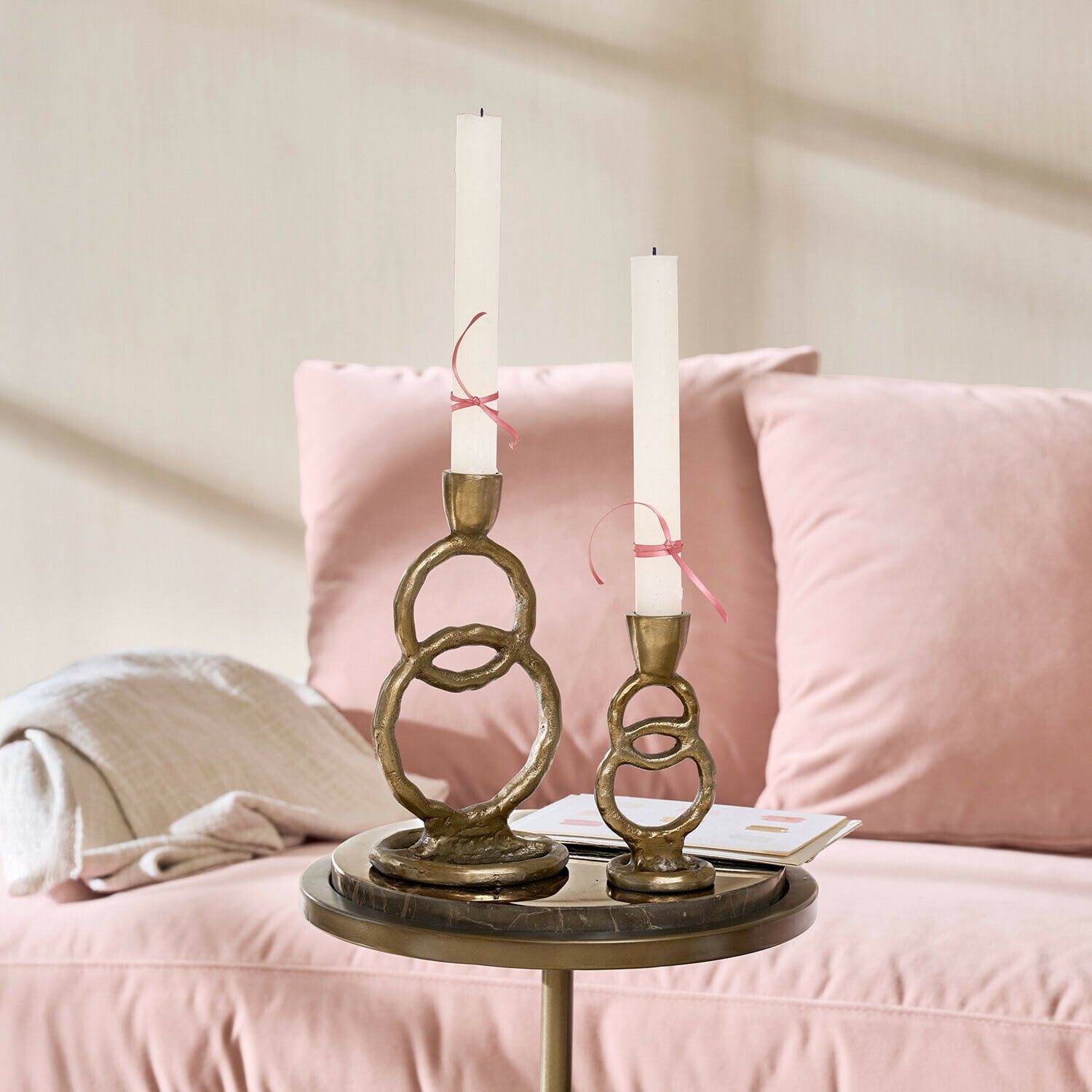 Kerzenständer 2er Set Corvane Set mit zwei messingfarbenen Kerzenhaltern und weißen Kerzen auf einem Tisch neben einem rosa Sofa