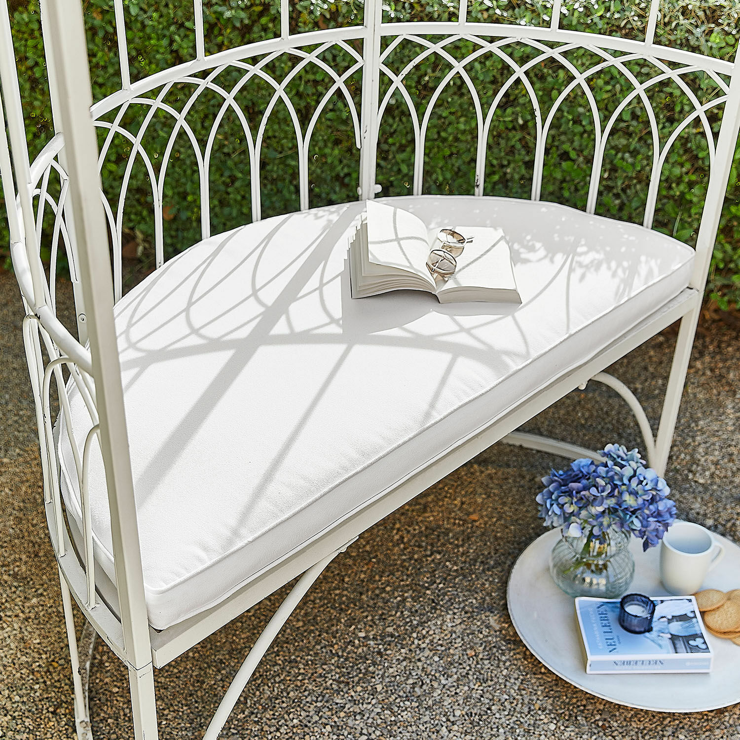 Sitzkissen Drivesto Weisse Metall-Gartenbank mit weissem Polster, offenem Buch, blauen Blumen und Tasse auf Terrasse