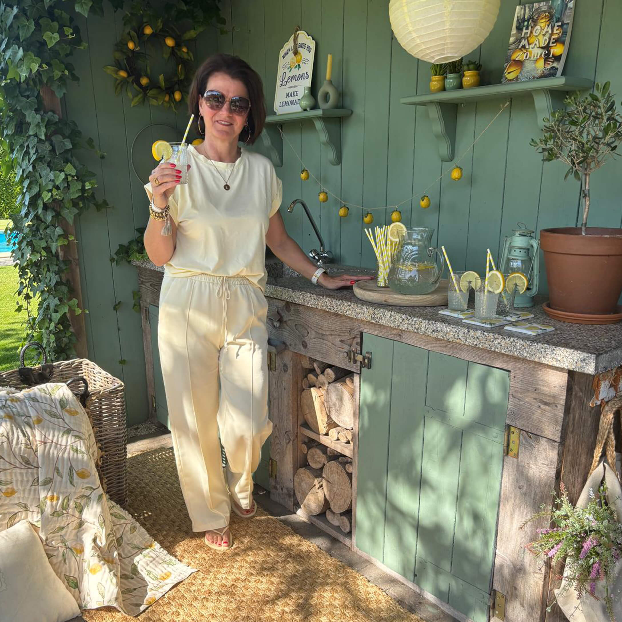 Frau im cremefarbenen Outfit steht an einer grünen Aussenküche mit Limonade und Zitronendekor.
