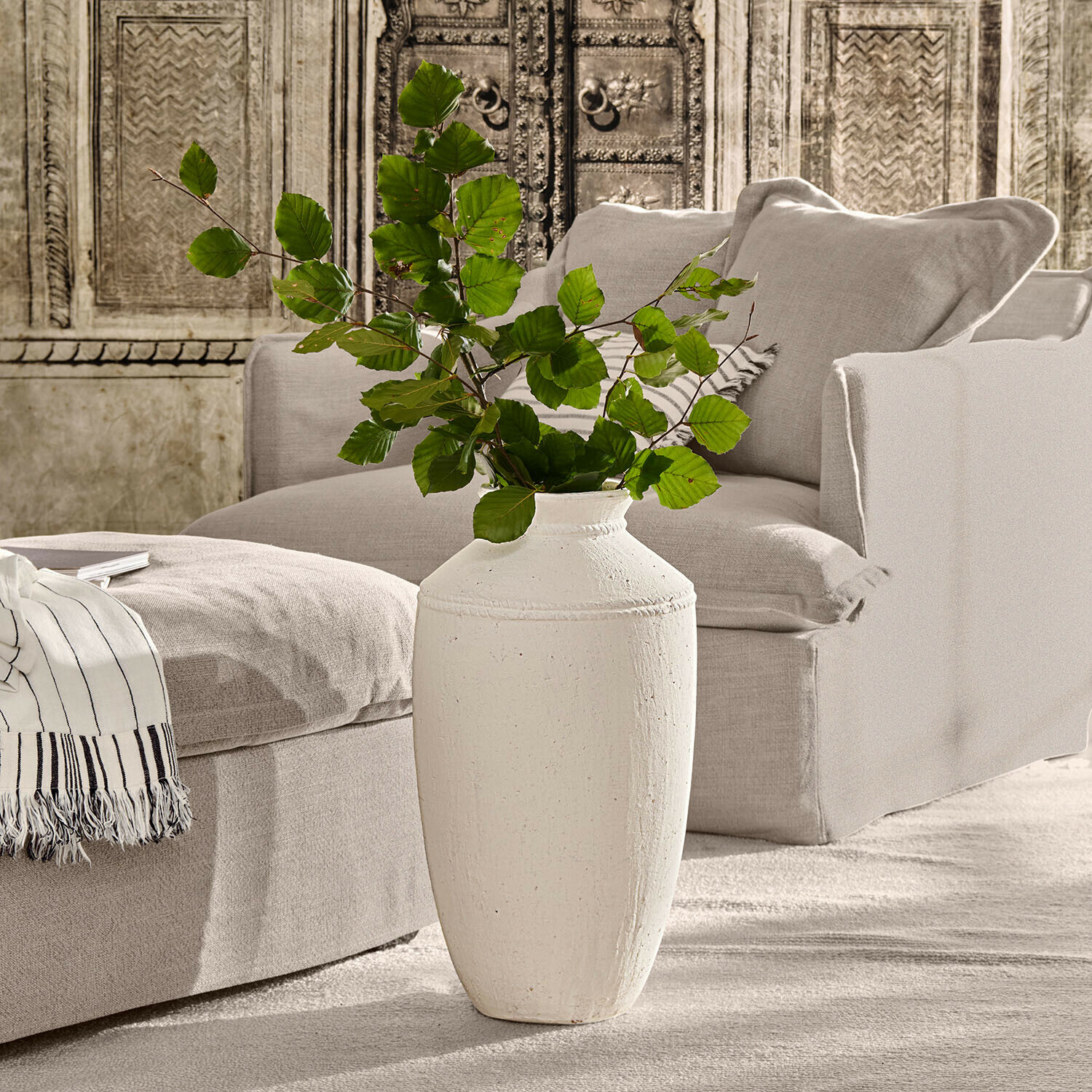 Weisse Keramikvase mit grünen Blättern vor beigem Sofa und dekorativer Wand im Wohnzimmer