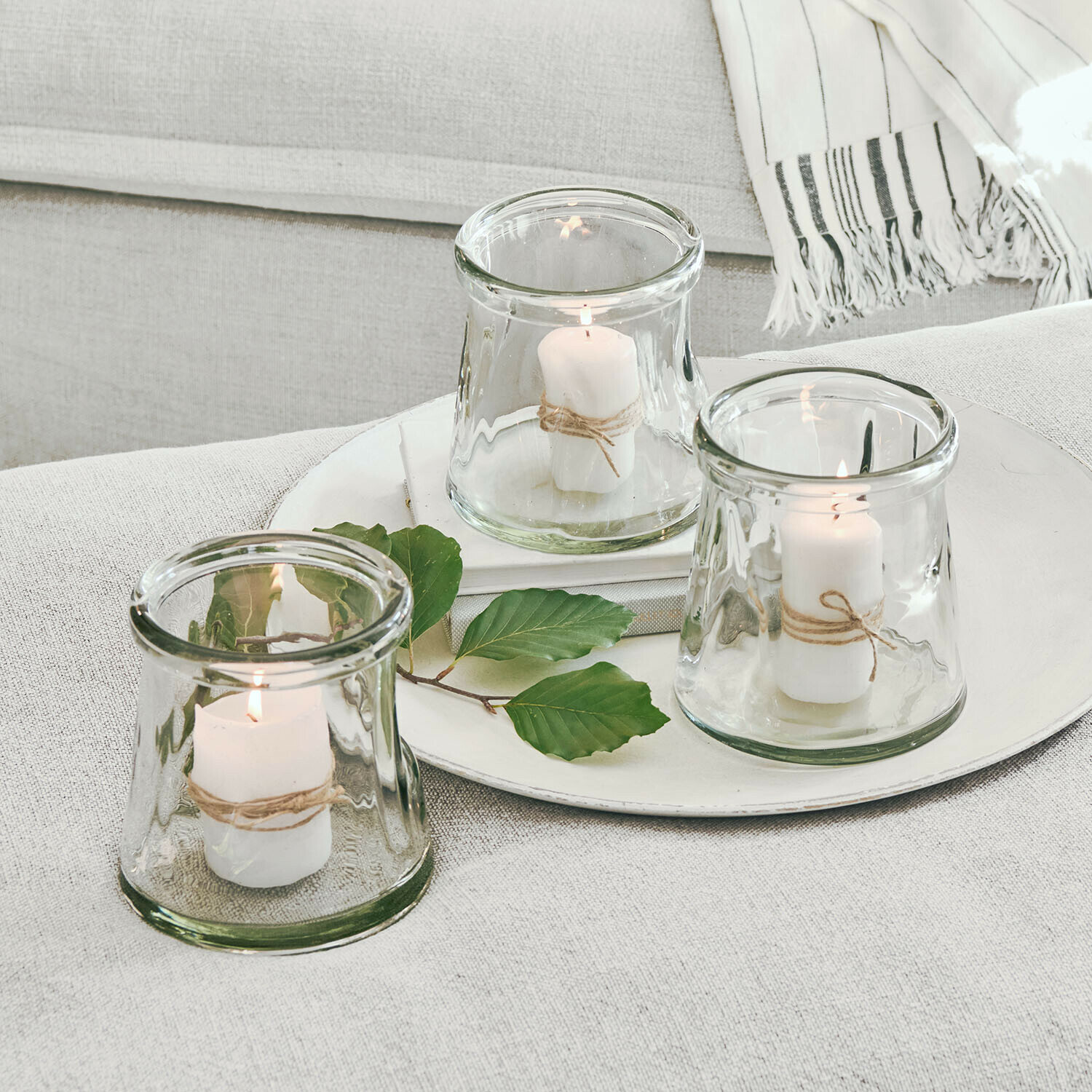 Set mit drei Glaswindlichtern mit weissen Kerzen und Naturkordel auf hellem Hintergrund