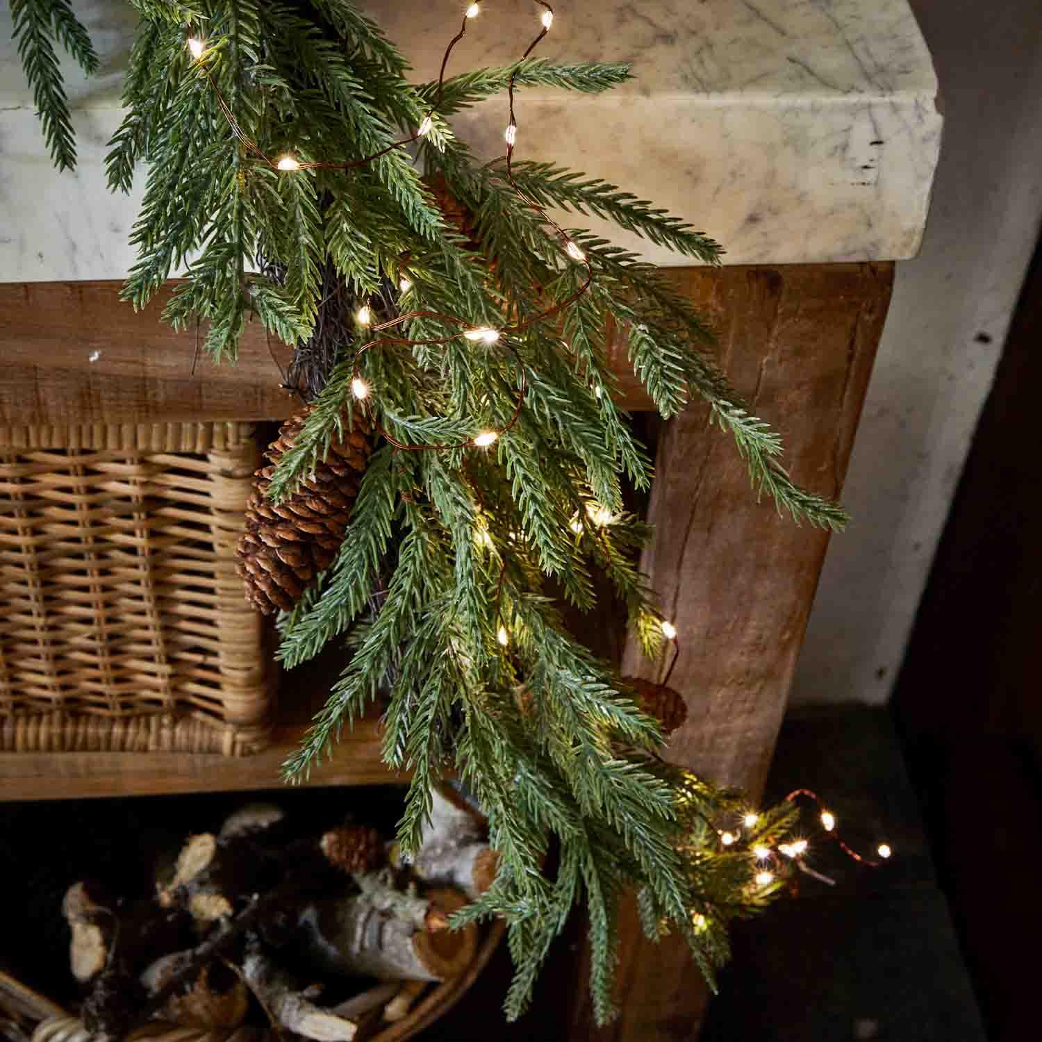 Tannenzweig mit Zapfen und LED-Lichterkette an Holzkamin, festliche Weihnachtsdekoration
