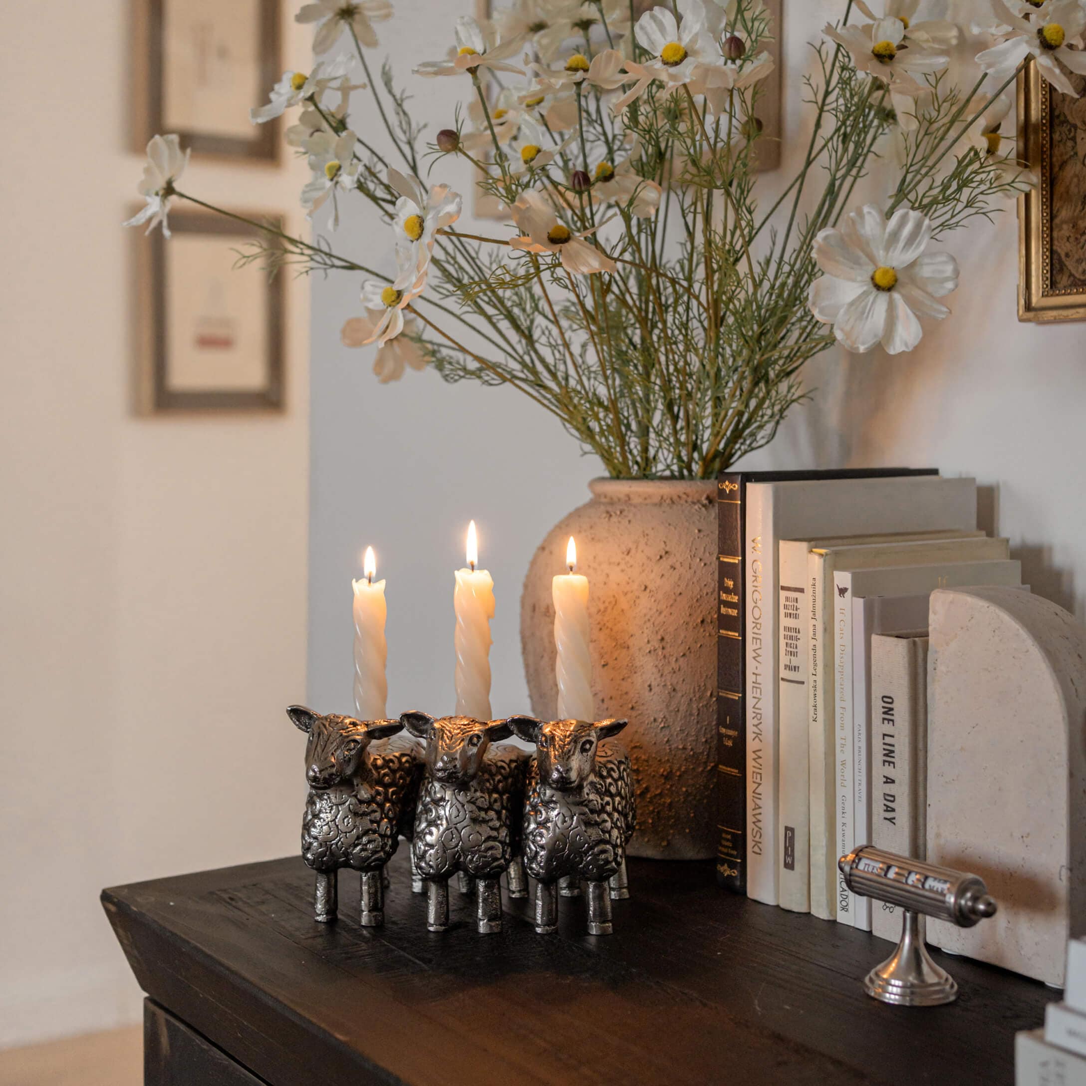 Metall-Kerzenhalter in Form von drei Schafen mit weissen Kerzen auf dunklem Holztisch neben Büchern