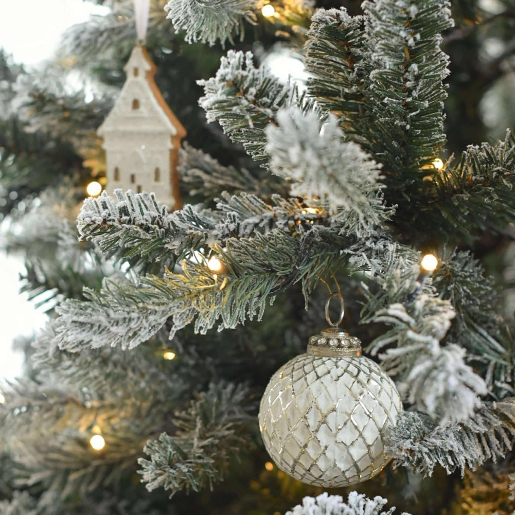 Weisse Glas-Weihnachtskugel mit Rautenmuster an schneebedecktem Tannenbaum mit LED-Lichtern und Holzdeko