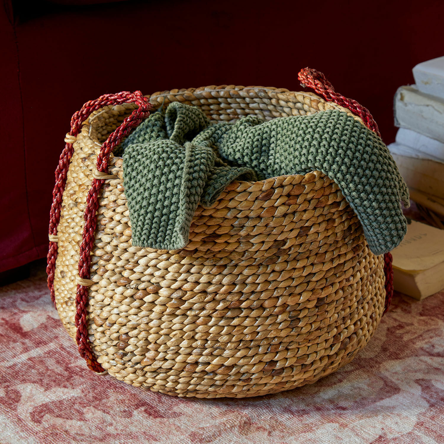 Korb Cilovar Runder Korb aus geflochtener Wasserhyazinthe mit roten Griffen und grünem Strickdecke auf Teppich