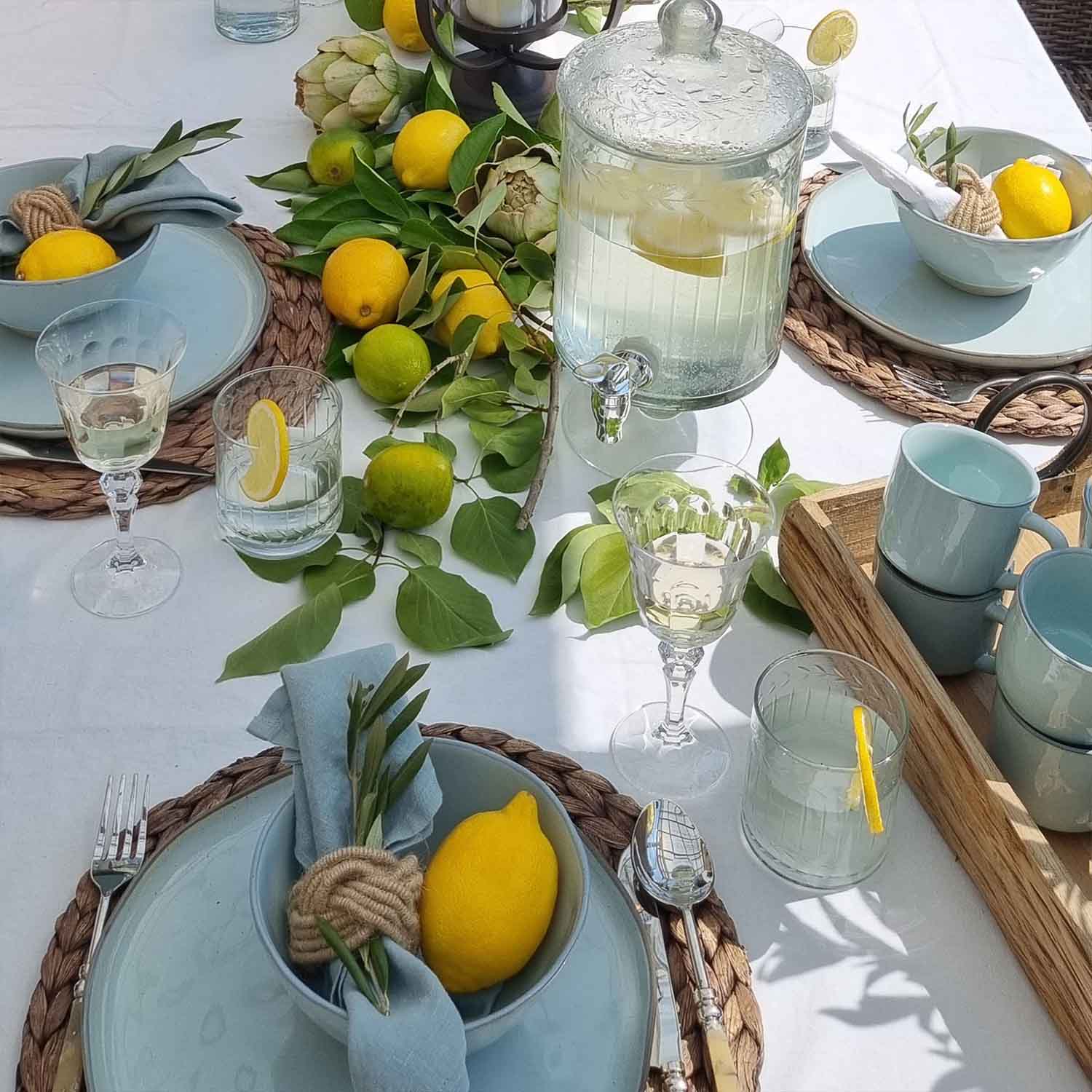 Sommerlicher Tisch mit blauer Keramik, Zitronen und grünen Blättern auf weissem Tischtuch, Tischdeko