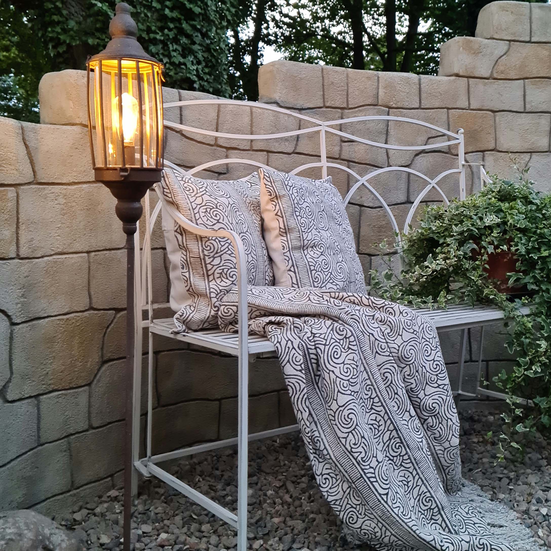 Weisse Metall-Gartenbank mit dekorativen Kissen und gemustertem Plaid, daneben eine beleuchtete Stehlampe