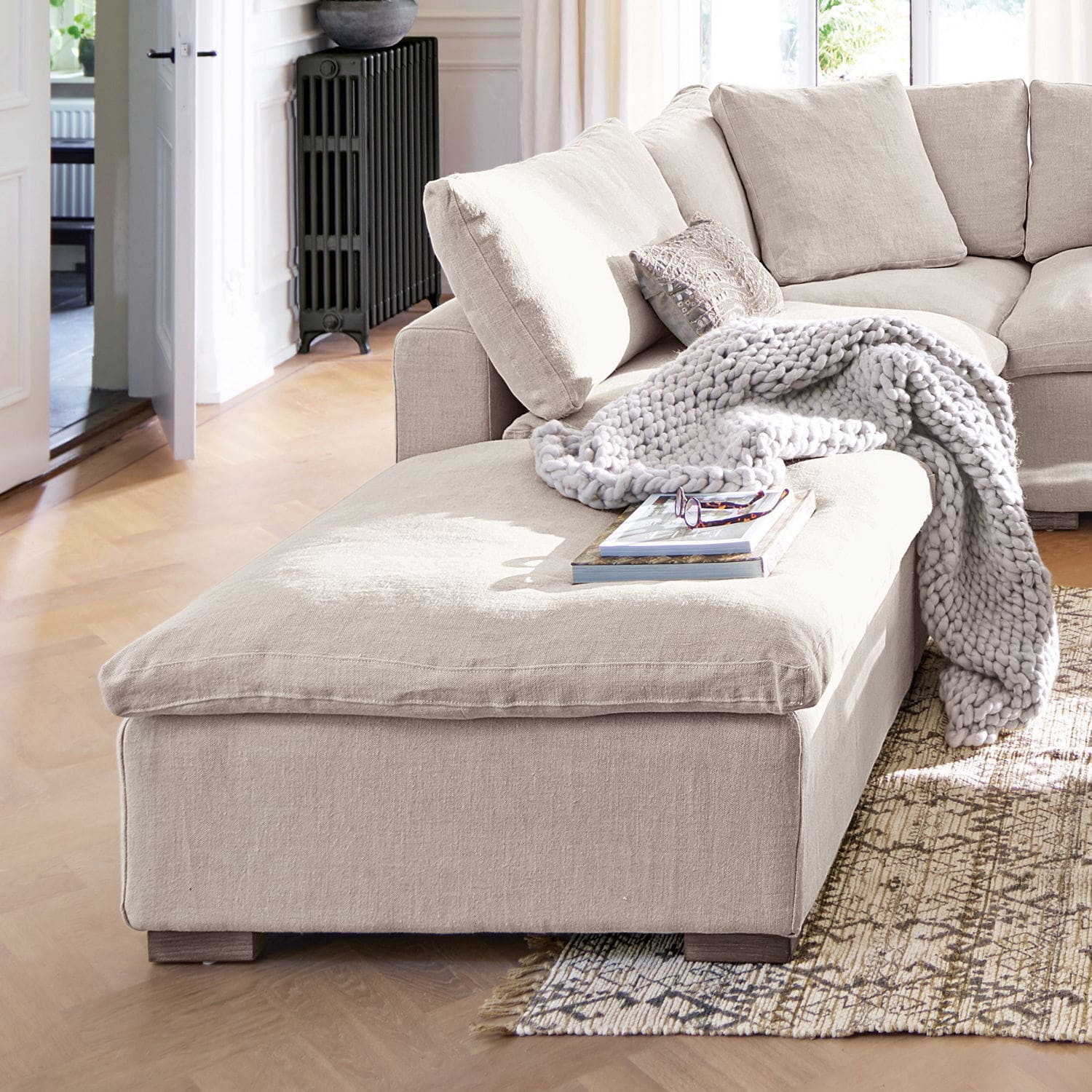 Hocker Bennington Beiges Ecksofa aus Stoff mit weicher Decke und Kissen in hellem Wohnzimmer mit Holzboden