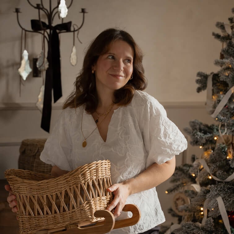 Frau in weisser bestickter Bluse hält einen Korb aus Weide auf Holzschlitten neben geschmücktem Weihnachtsbaum
