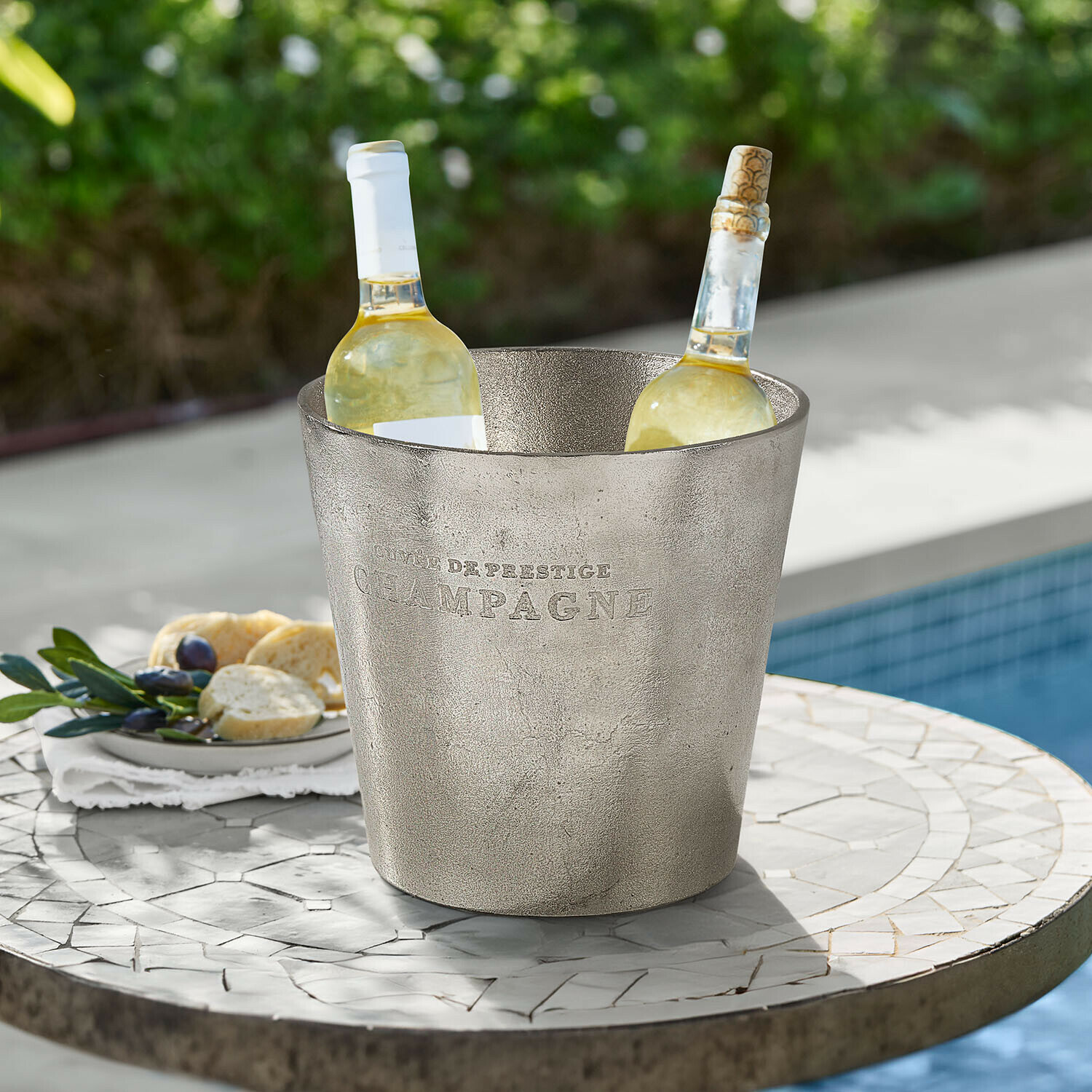 Silberner Metall-Eiskübel mit Aufschrift Champagne und zwei Weissweinflaschen auf Gartentisch