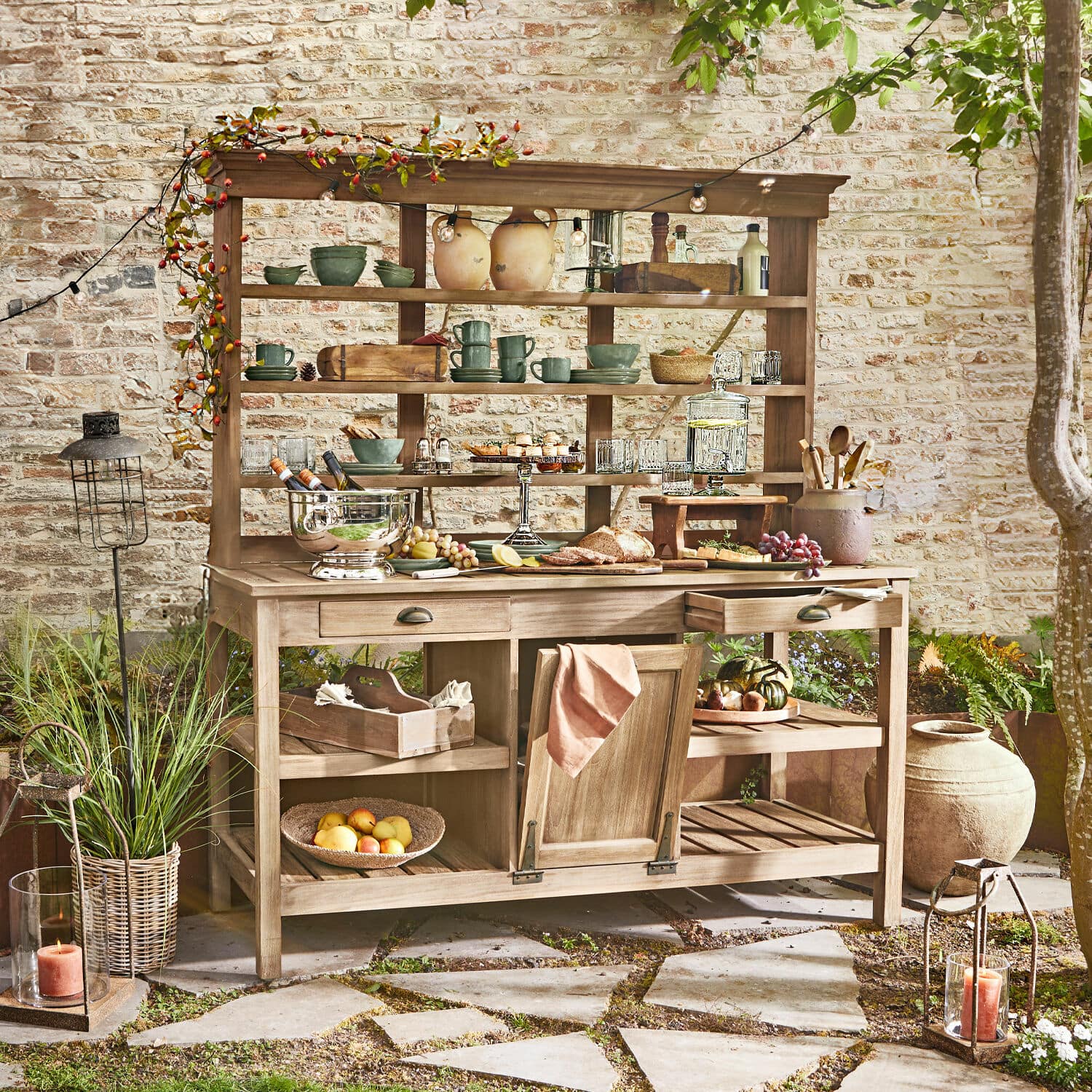 Gartenschrank Fallon Holz-Gartenmöbel mit Regalen und Schubladen, dekoriert mit Geschirr und Speisen auf der Terrasse