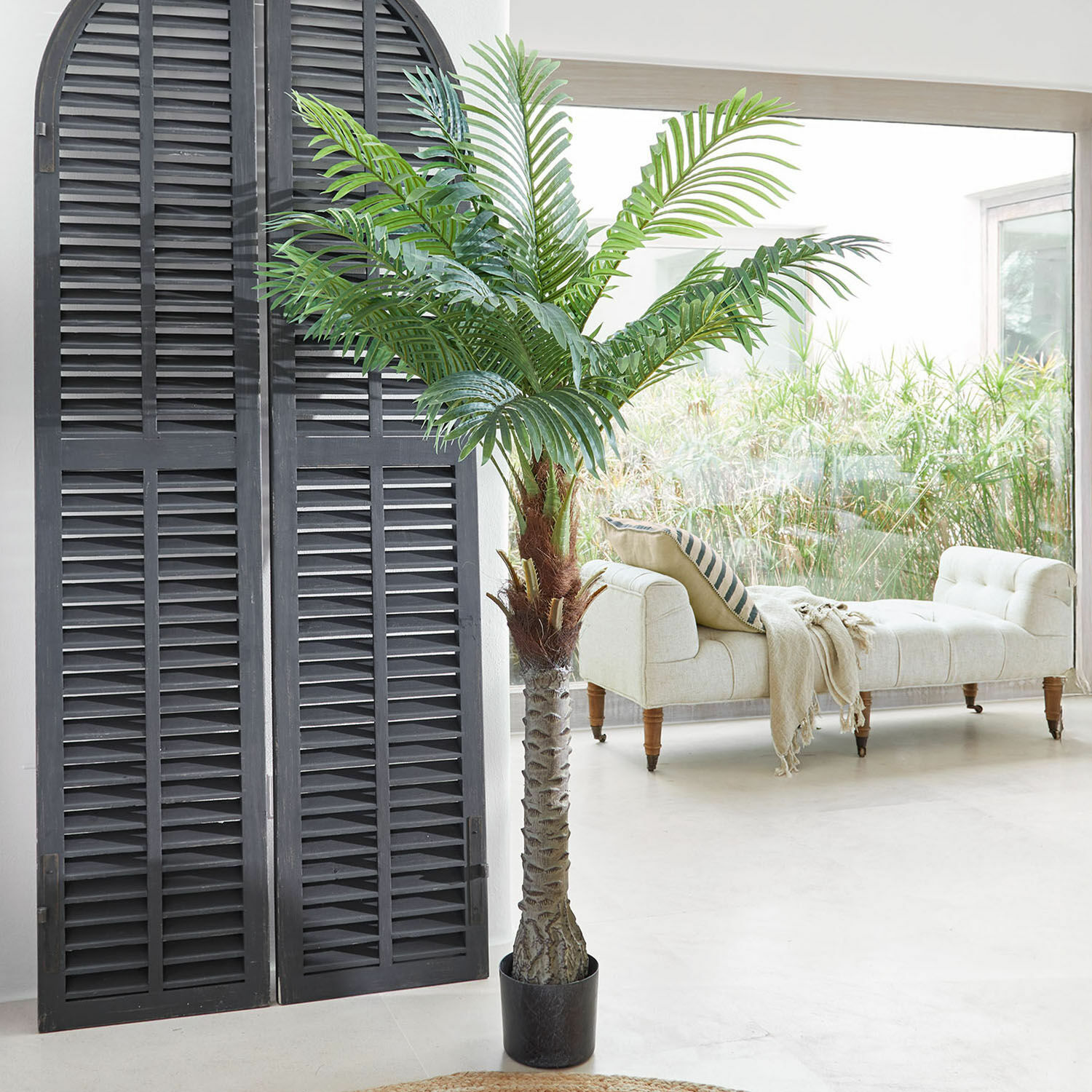 Deko-Topfpflanze Zephyriel Künstliche Palme in schwarzem Topf im hellen Wohnzimmer mit beigem Sofa und grossem Fenster