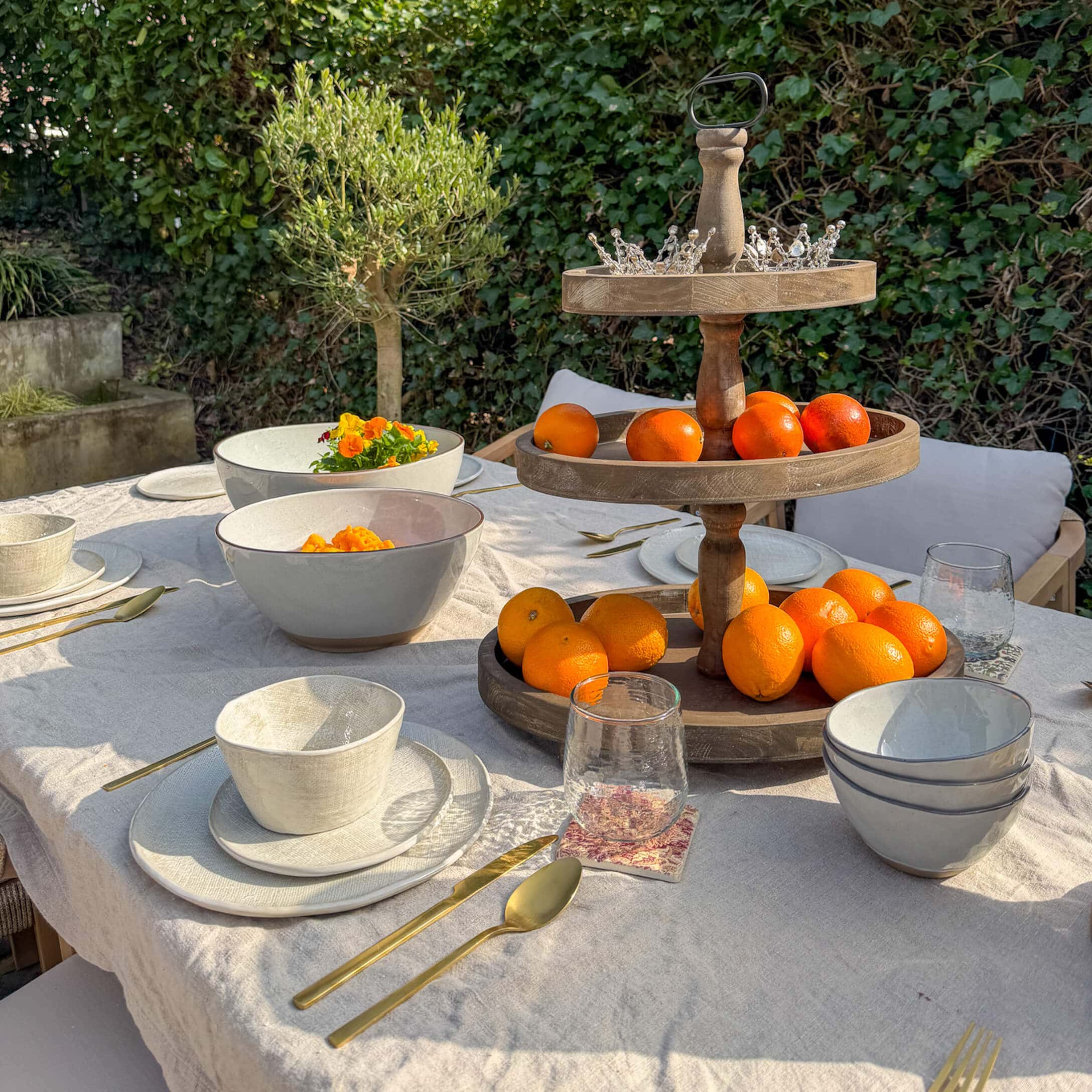 Gartentisch mit weissem Keramikgeschirr, goldenen Bestecken und hölzernem Obstständer mit Orangen