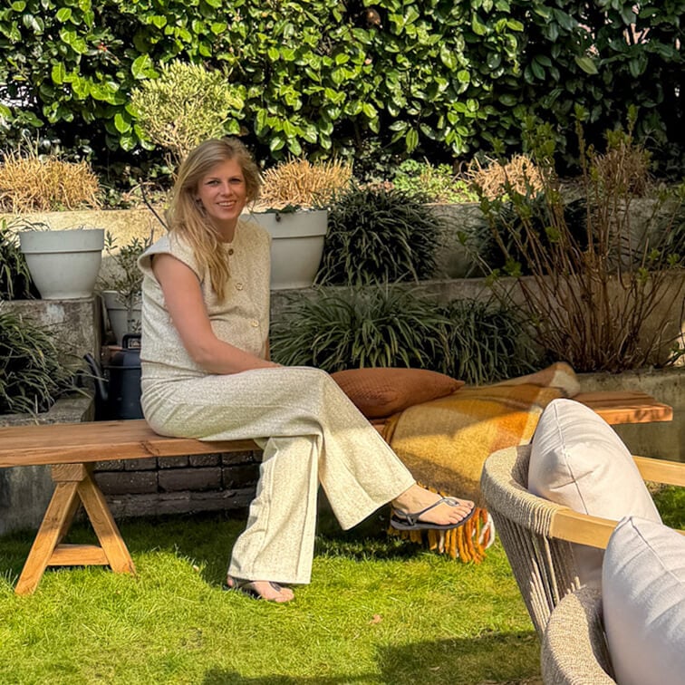 Frau in hellem Outfit sitzt auf Holzbank im Garten mit grünen Pflanzen und Kissen