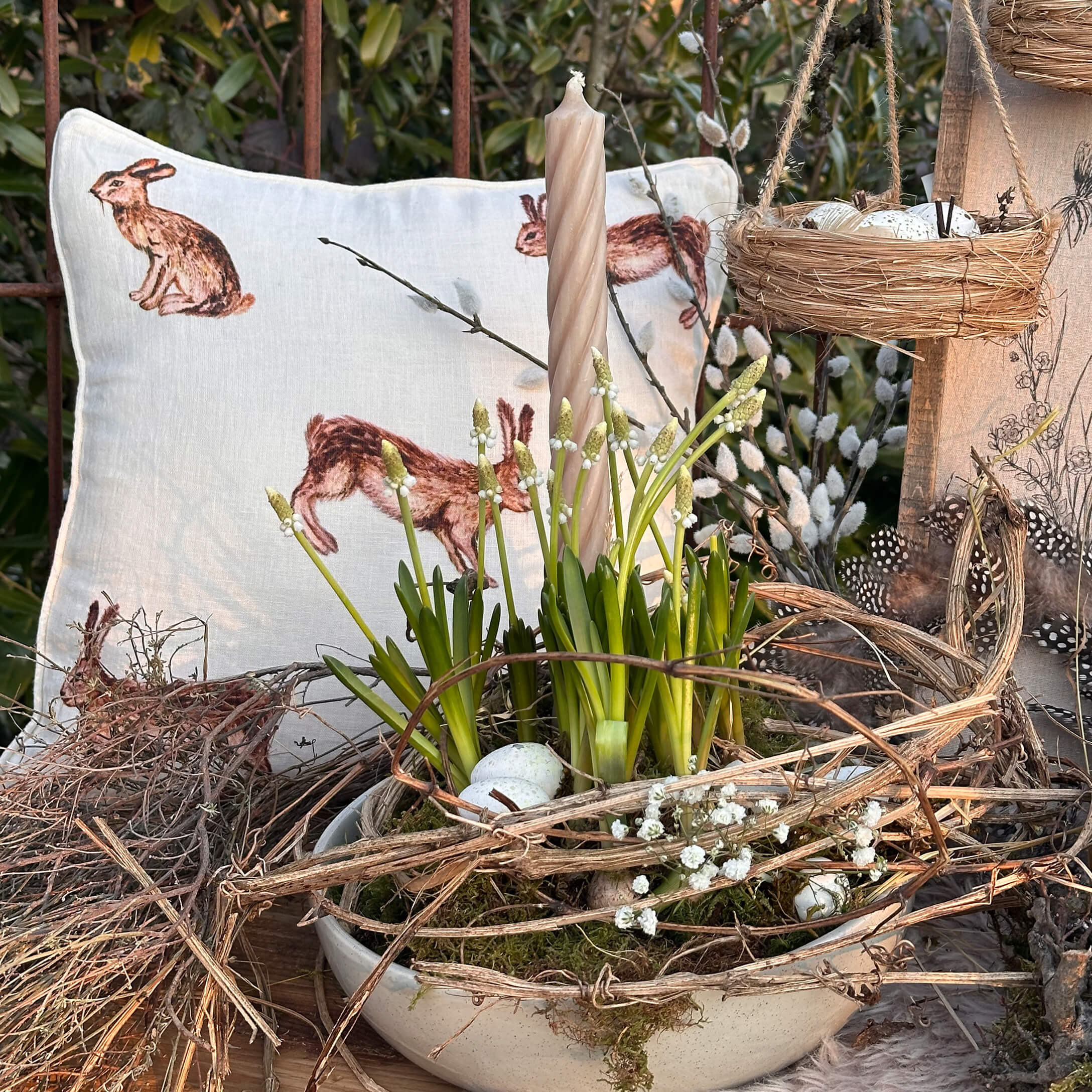 Natürlich dekorierte Osterfreude mit weisser Kerze, grünen Blumen und Hasenmotiv-Kissen auf Holztisch