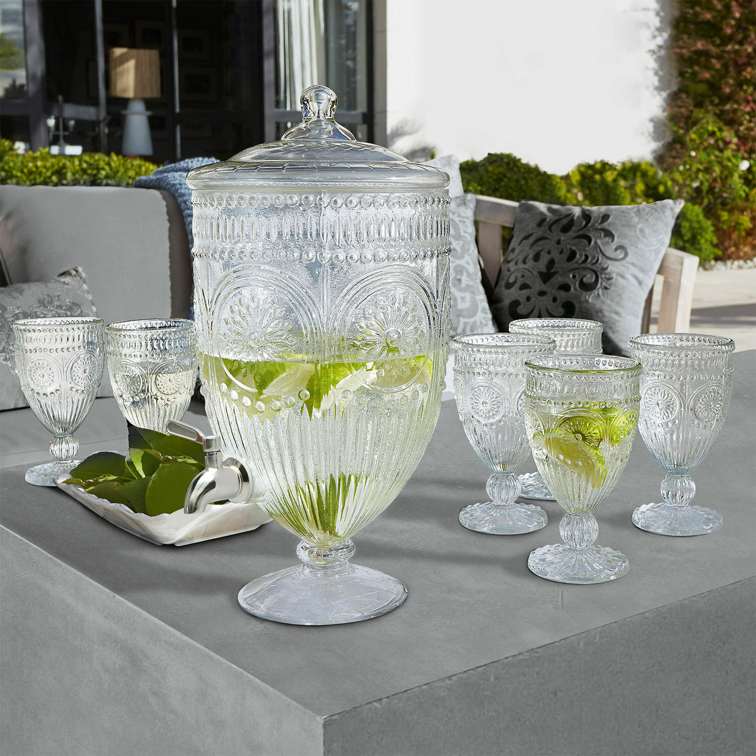 Glas-Getränkespender mit Deckel und sechs verzierten Gläsern auf Gartentisch